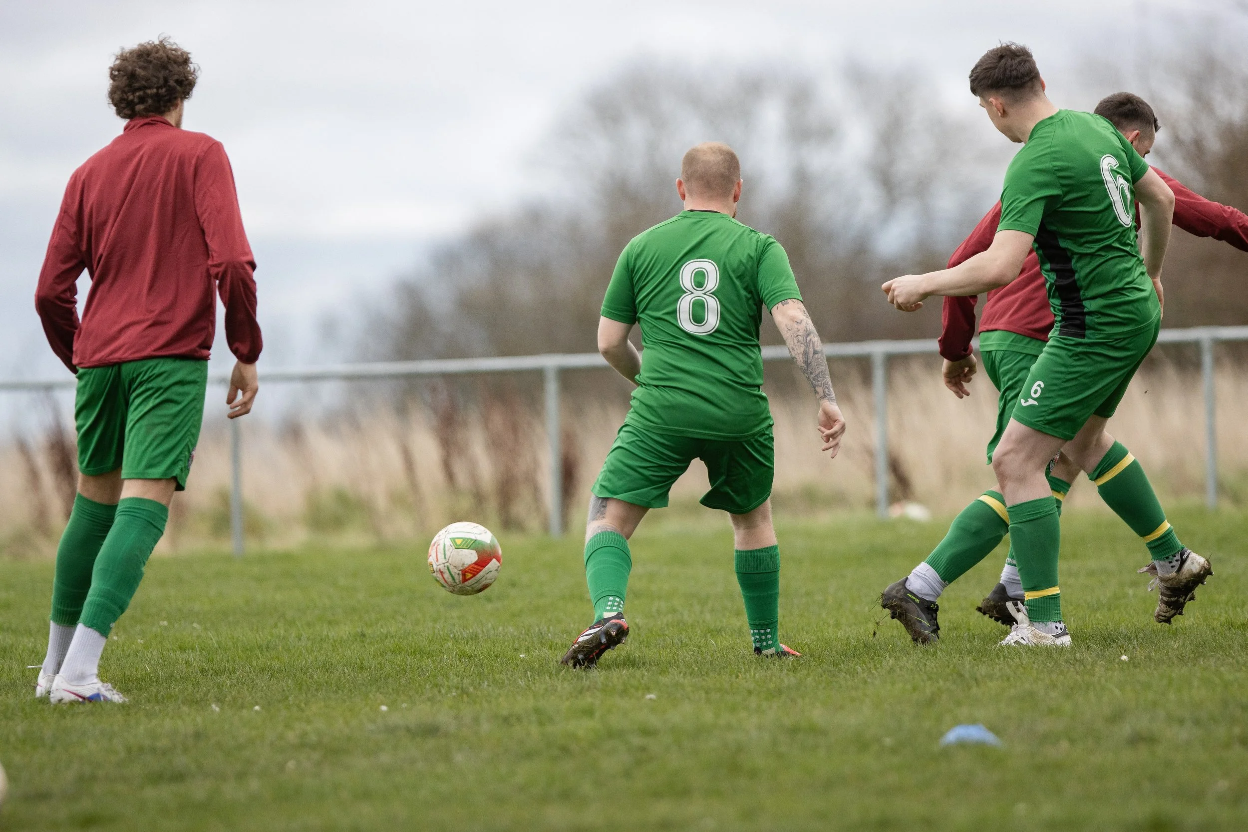 Soccer players practicing on the field during daytime, wearing green shorts, socks, and jerseys with numbers, with one in a goalkeeper uniform.