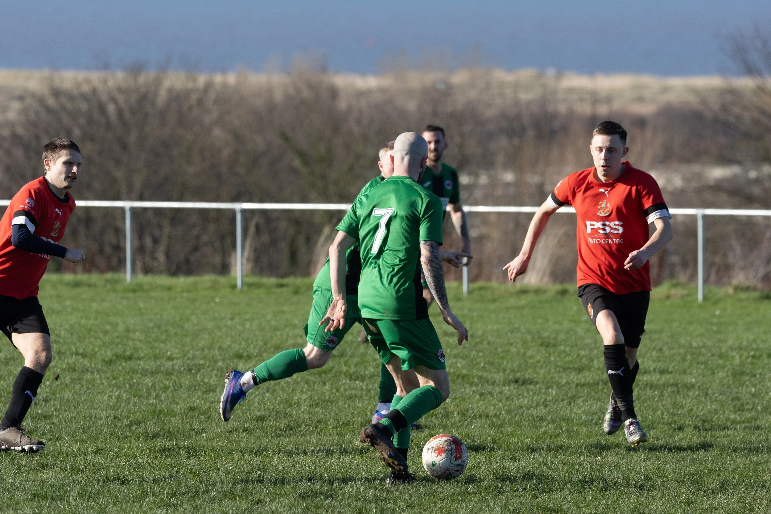 Soccer match with players in green and red jerseys on a grassy field, with a background of trees and hills.