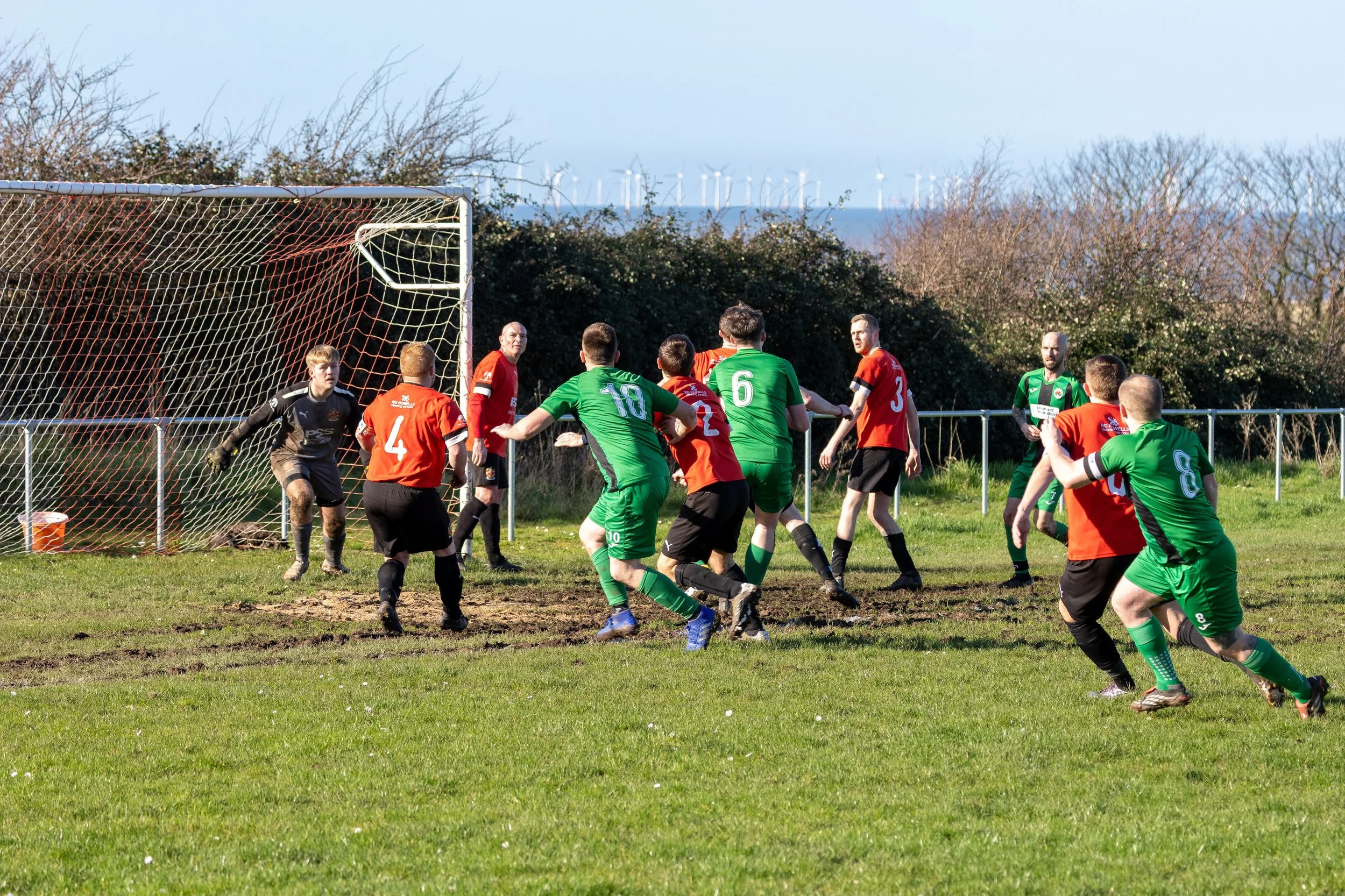 Soccer match in progress with players in red and green jerseys near the goal, goalkeeper ready, on a grassy field with trees and wind turbines in the background.