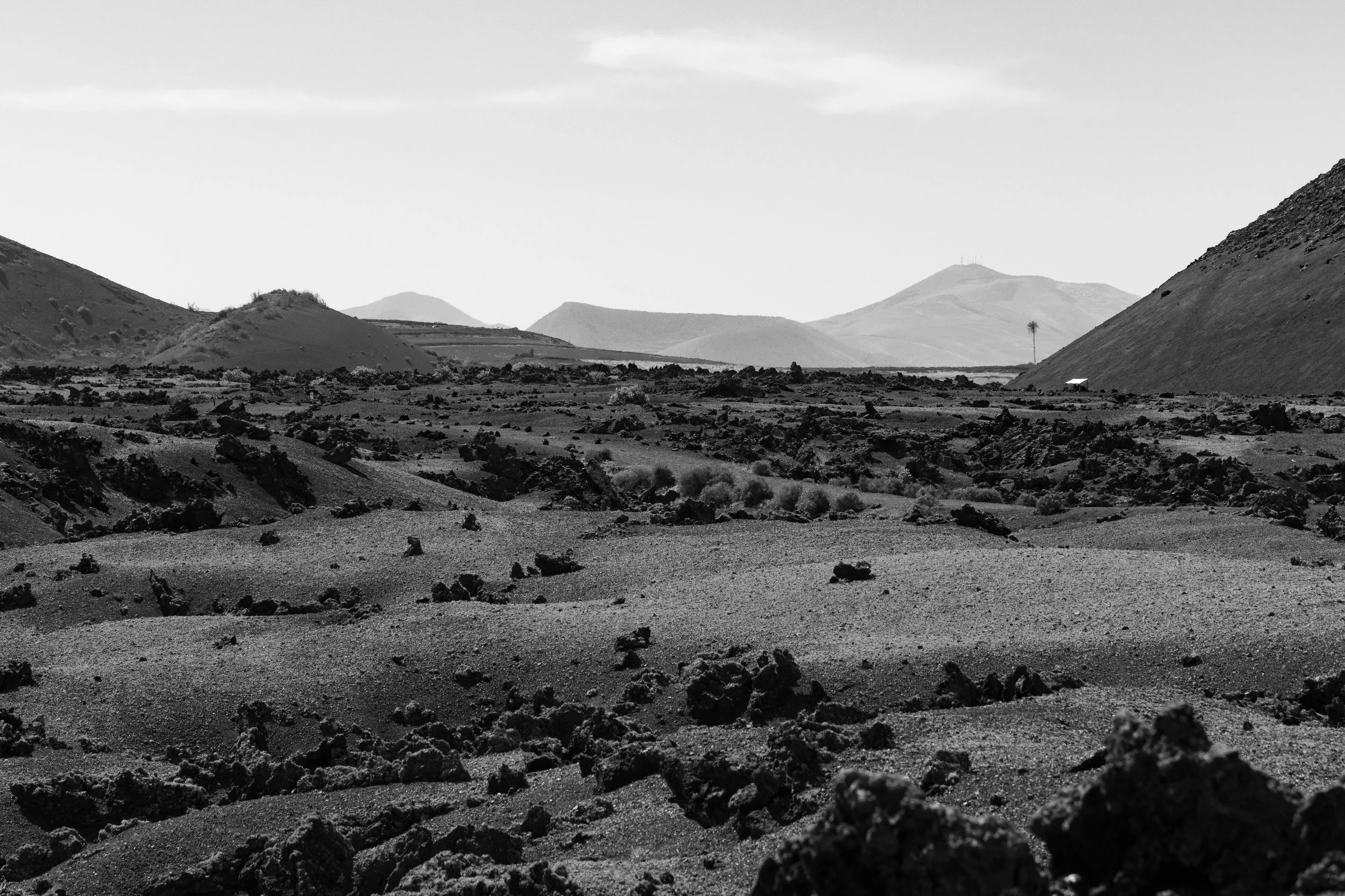 A black and white landscape photograph of a volcanic or rocky desert with distant hills and mountains in the background.