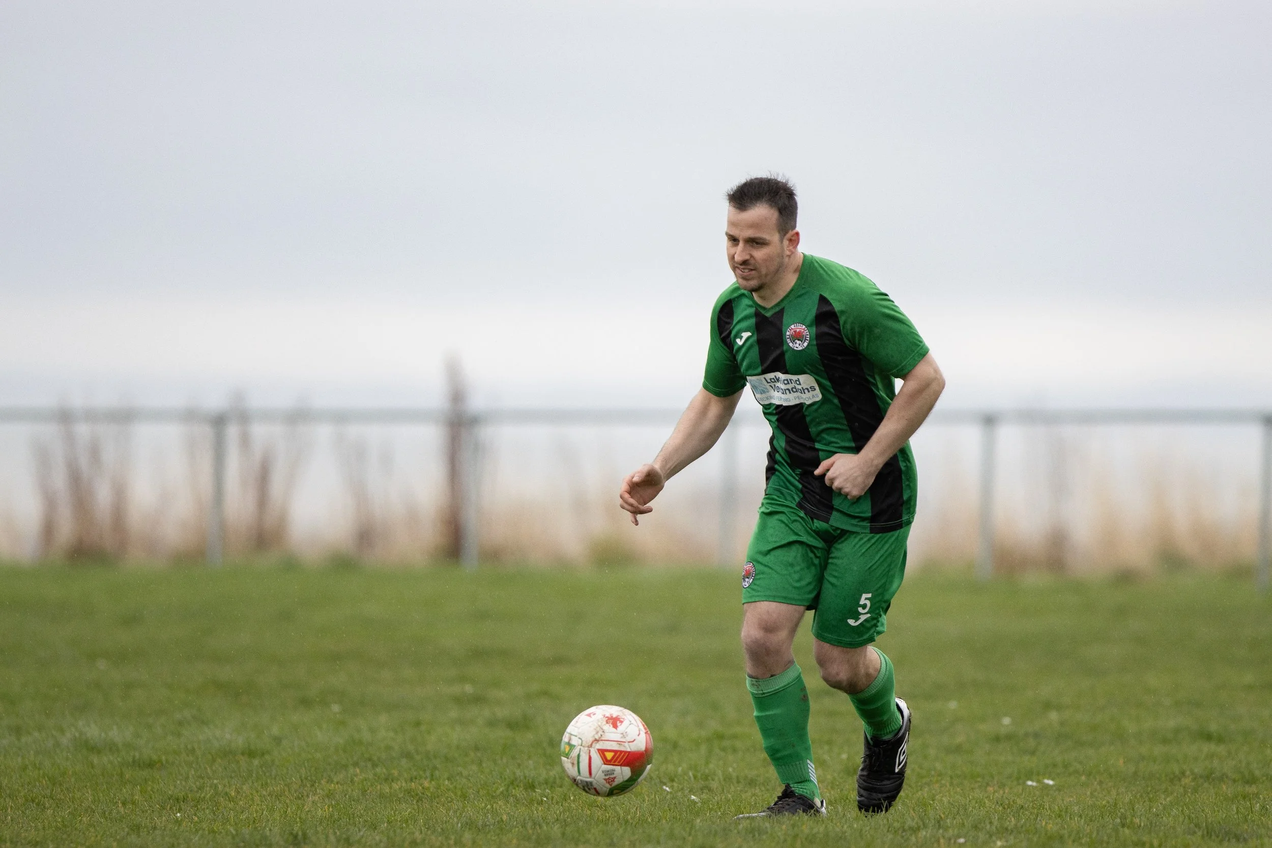 A man in a green and black soccer uniform controls a soccer ball on a grassy field with a cloudy sky in the background.