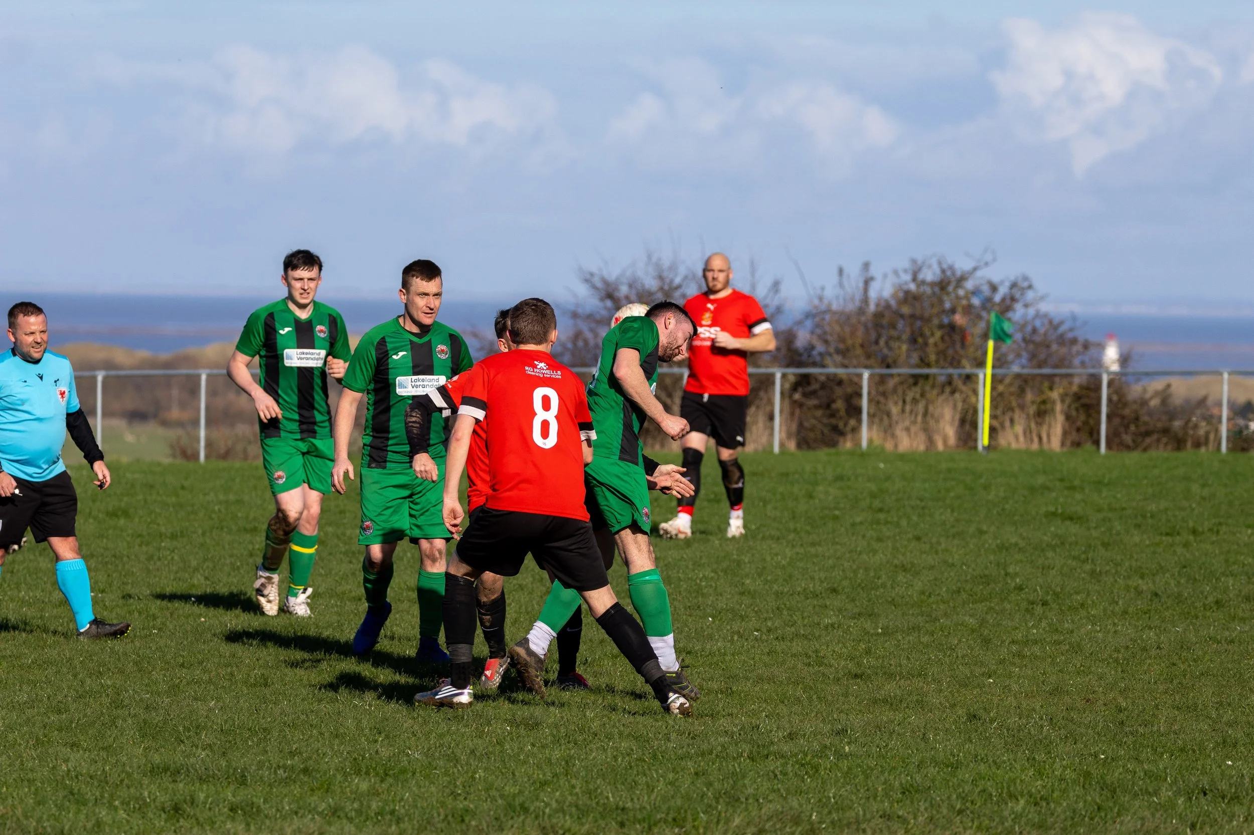 Soccer match with players in green and red jerseys on a grassy field, with a fence and trees in the background under a partly cloudy sky.