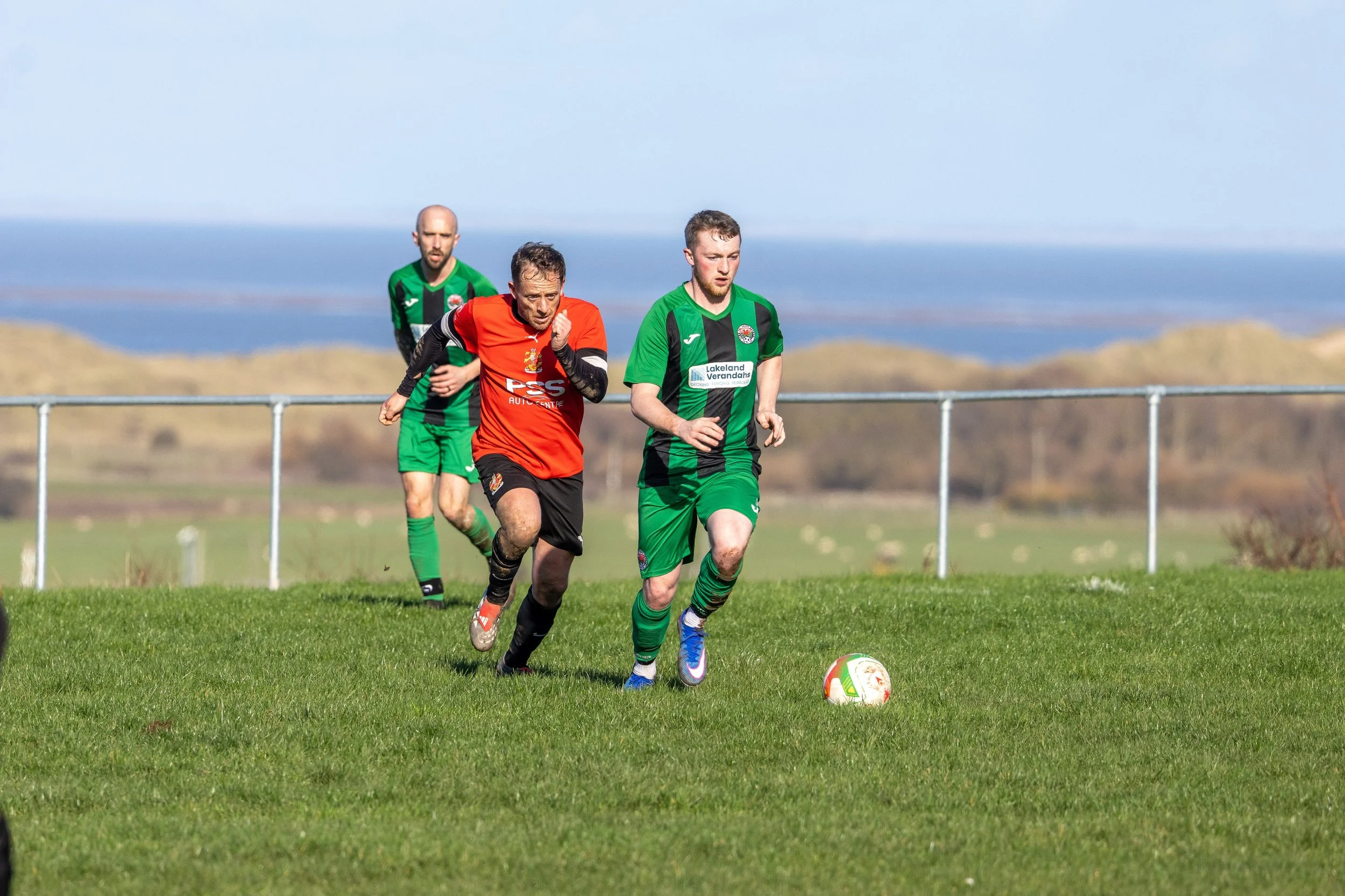 Three soccer players playing on a grassy field with a scenic background of hills and water. Two players are in green and black jerseys, and one player is in a red jersey, all running toward a soccer ball.
