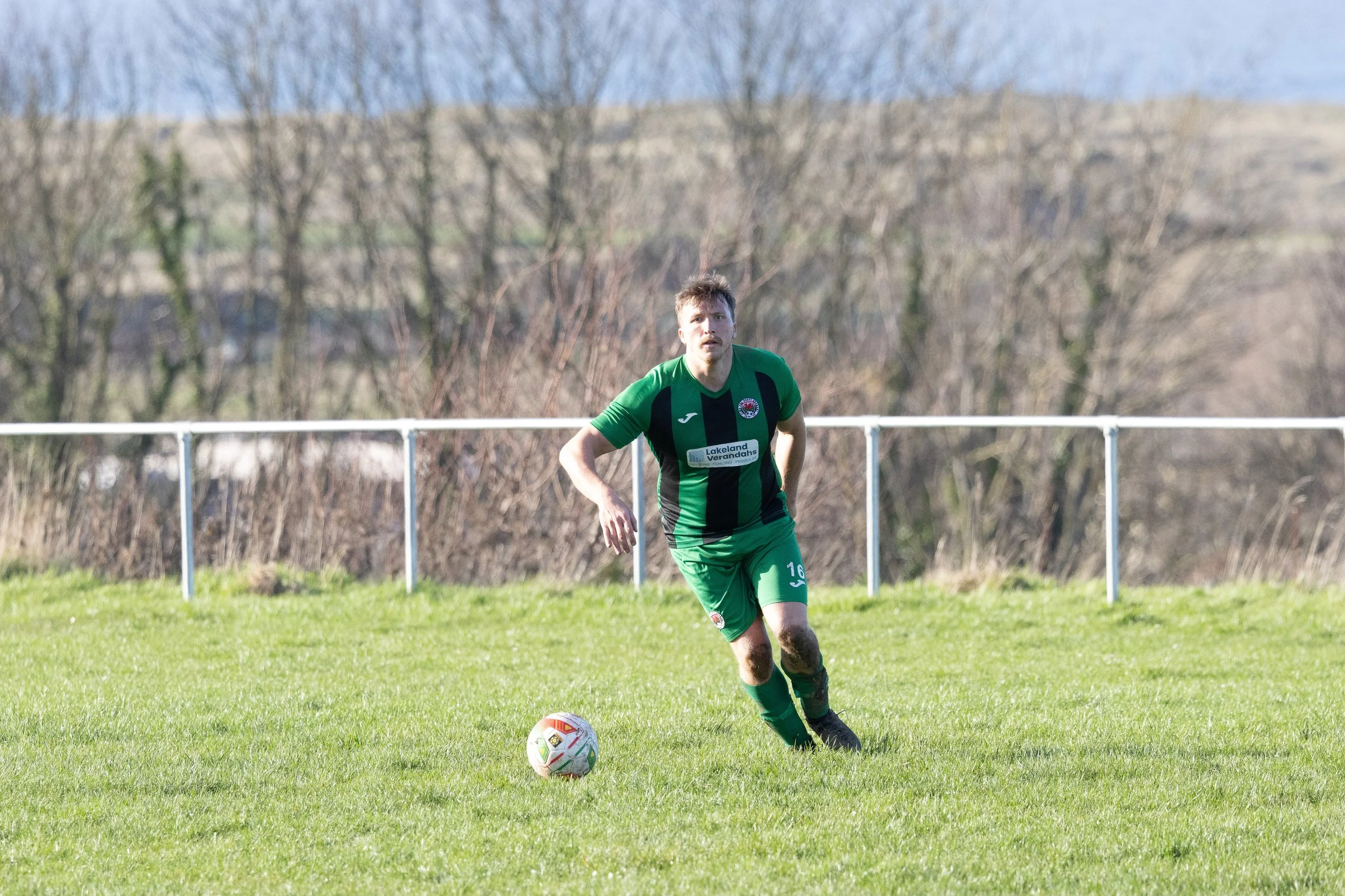 A soccer player in a green and black uniform running on a grassy field with a soccer ball nearby, during daytime.