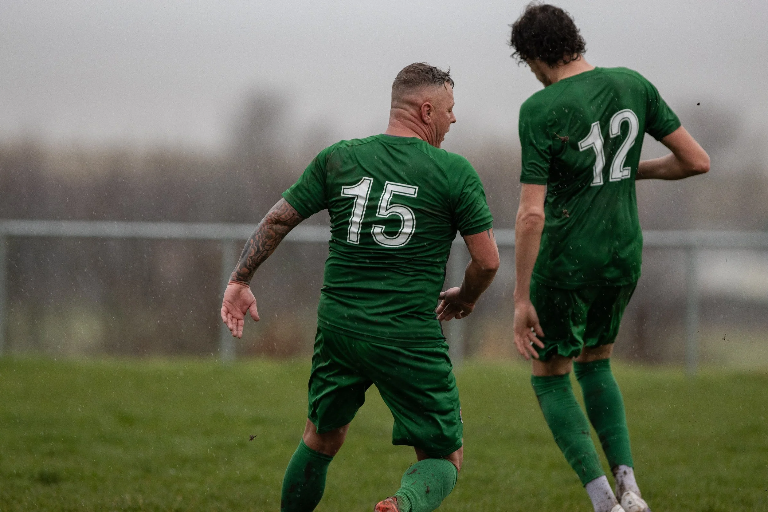 Two soccer players in green uniforms, numbers 15 and 12, standing on a muddy field in rainy weather, with blurred background.