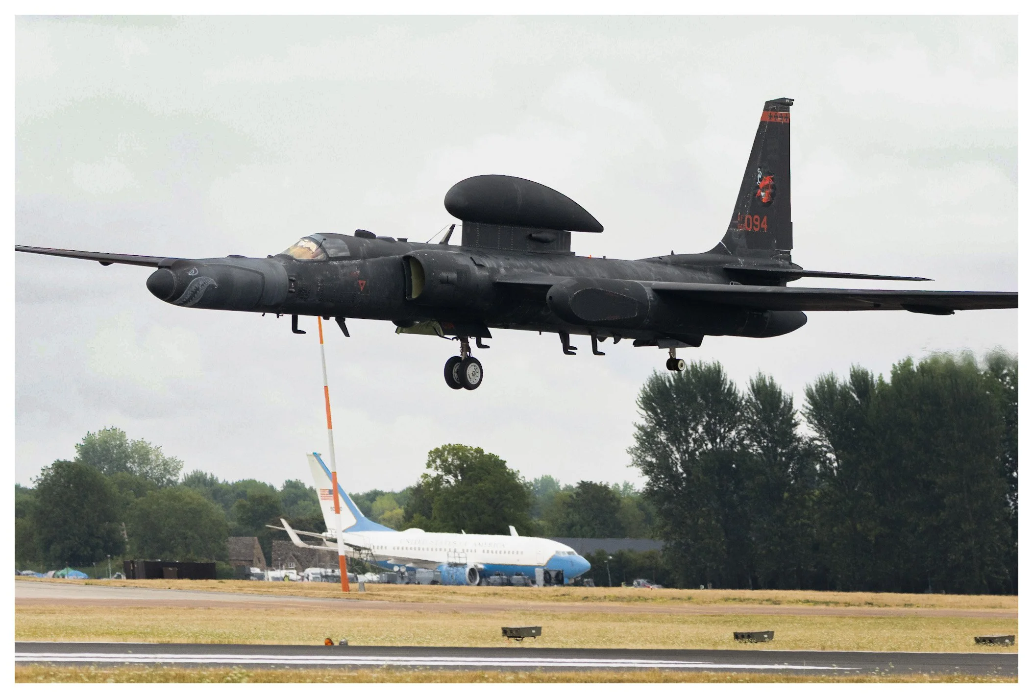 Black military jet aircraft flying low over an airfield with a commercial airplane parked on the tarmac and trees in the background.