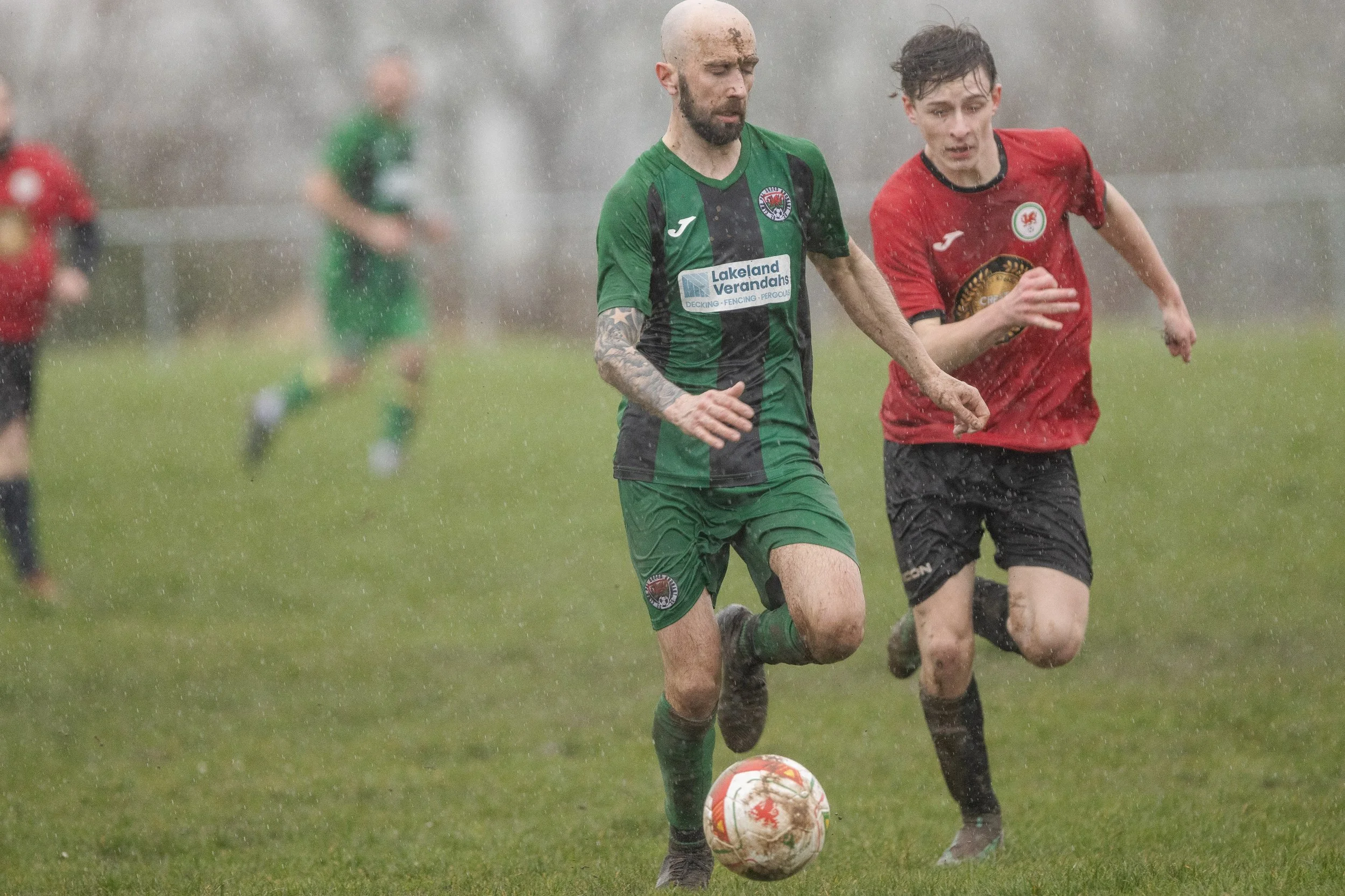 Two soccer players running in the rain on a grassy field, one in a green uniform and the other in a red uniform, with a soccer ball at their feet.