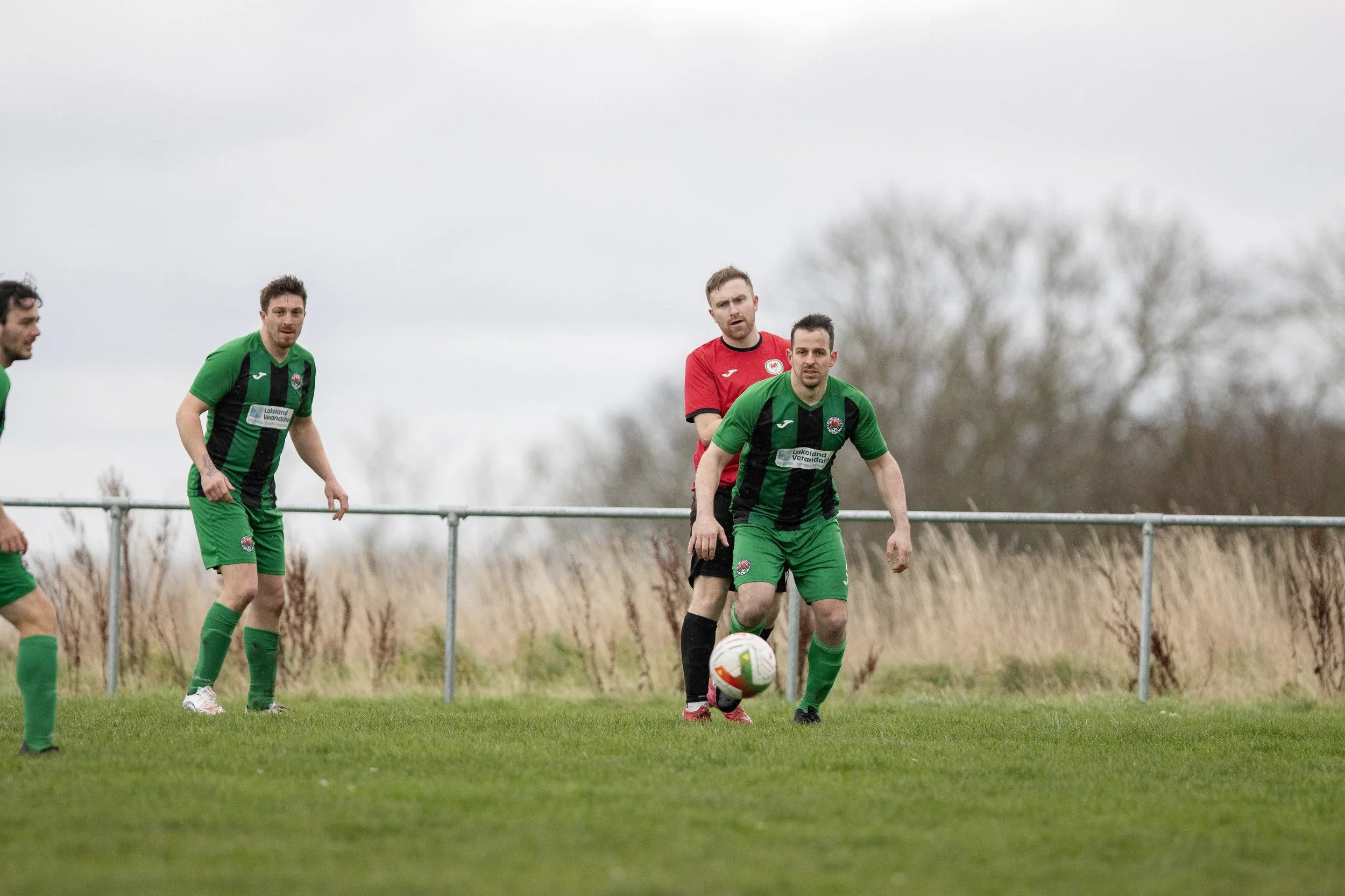 Soccer players on a grassy field, with some wearing green uniforms and one in a red uniform, during a match on a cloudy day.