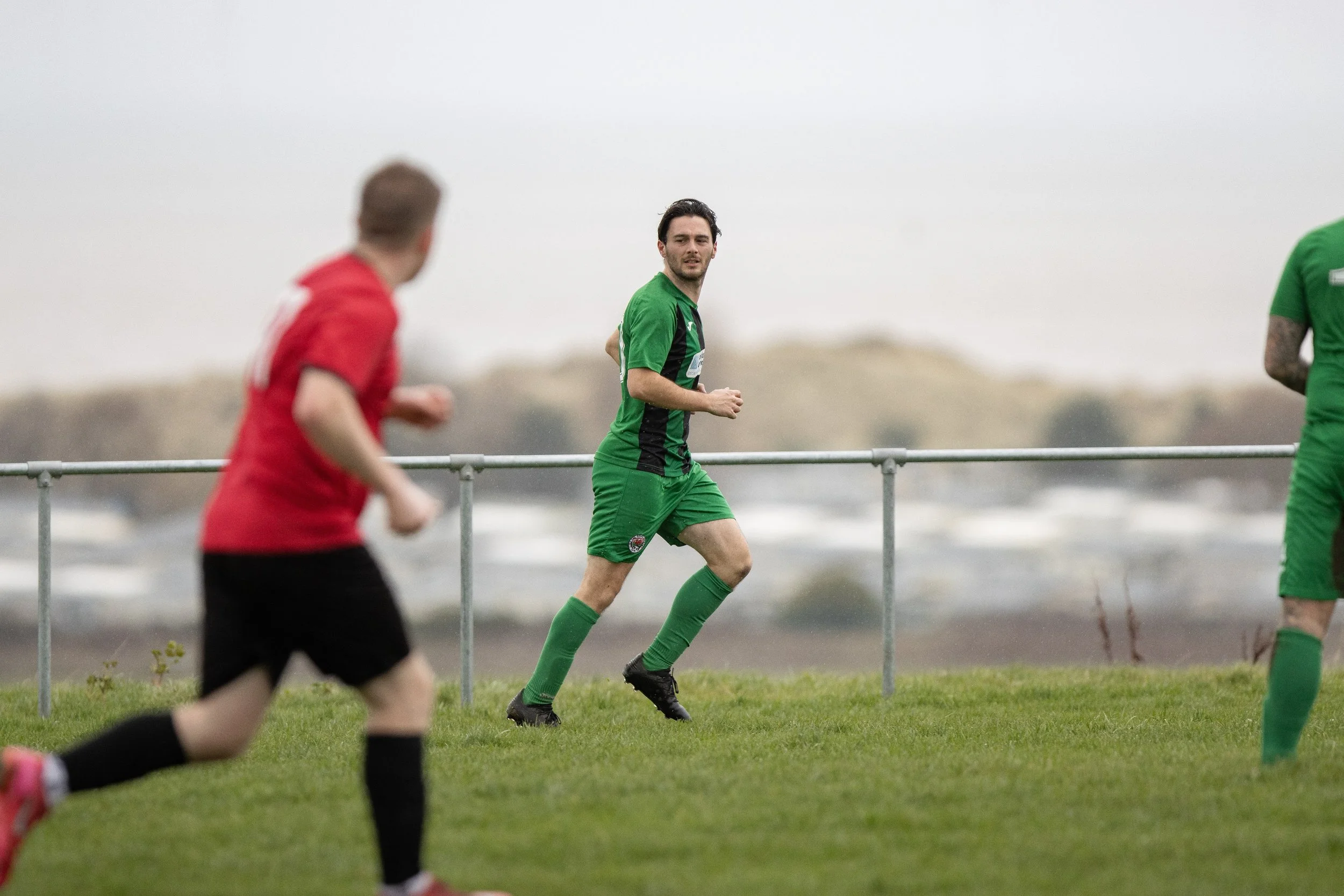 Soccer players on the field during a game, with one player in a green uniform looking towards the camera, and others in red and green uniforms running.