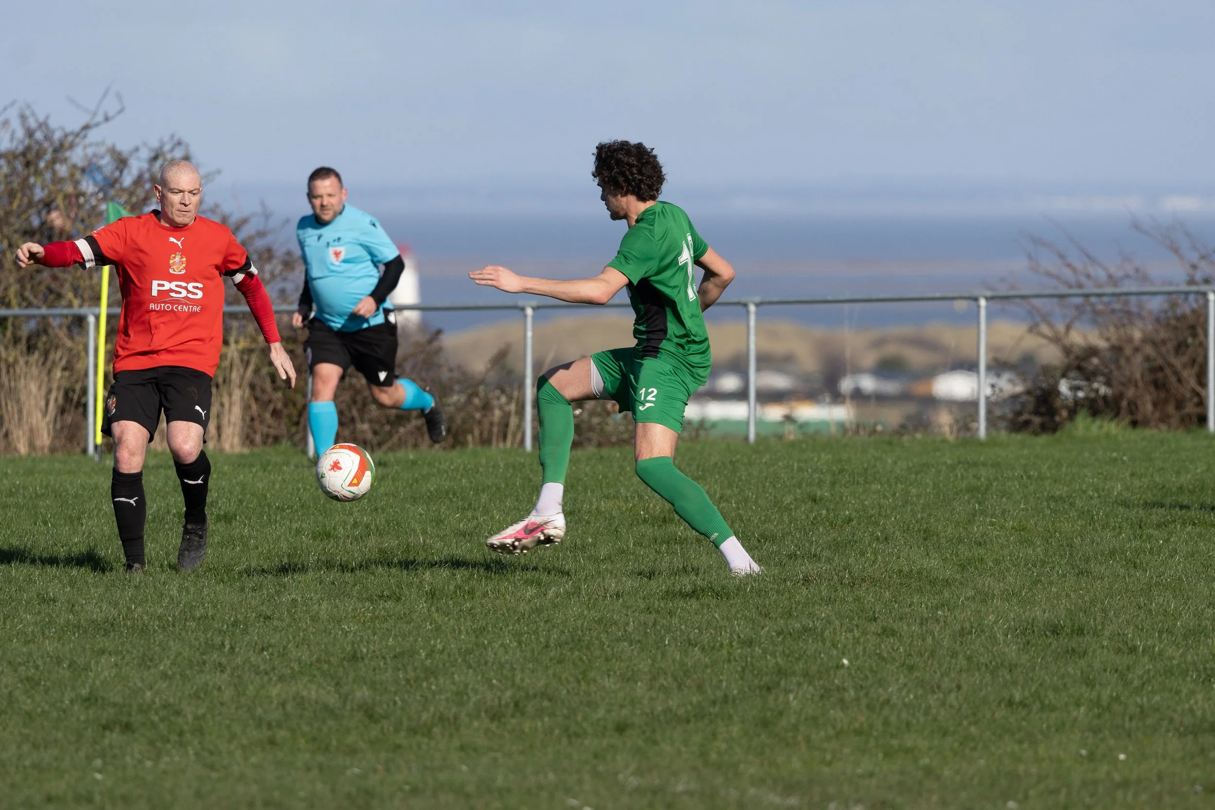Three soccer players in action on a grassy field under a cloudy sky. One player in green is trying to kick the ball, while two players, one in red and another in blue, are approaching him.