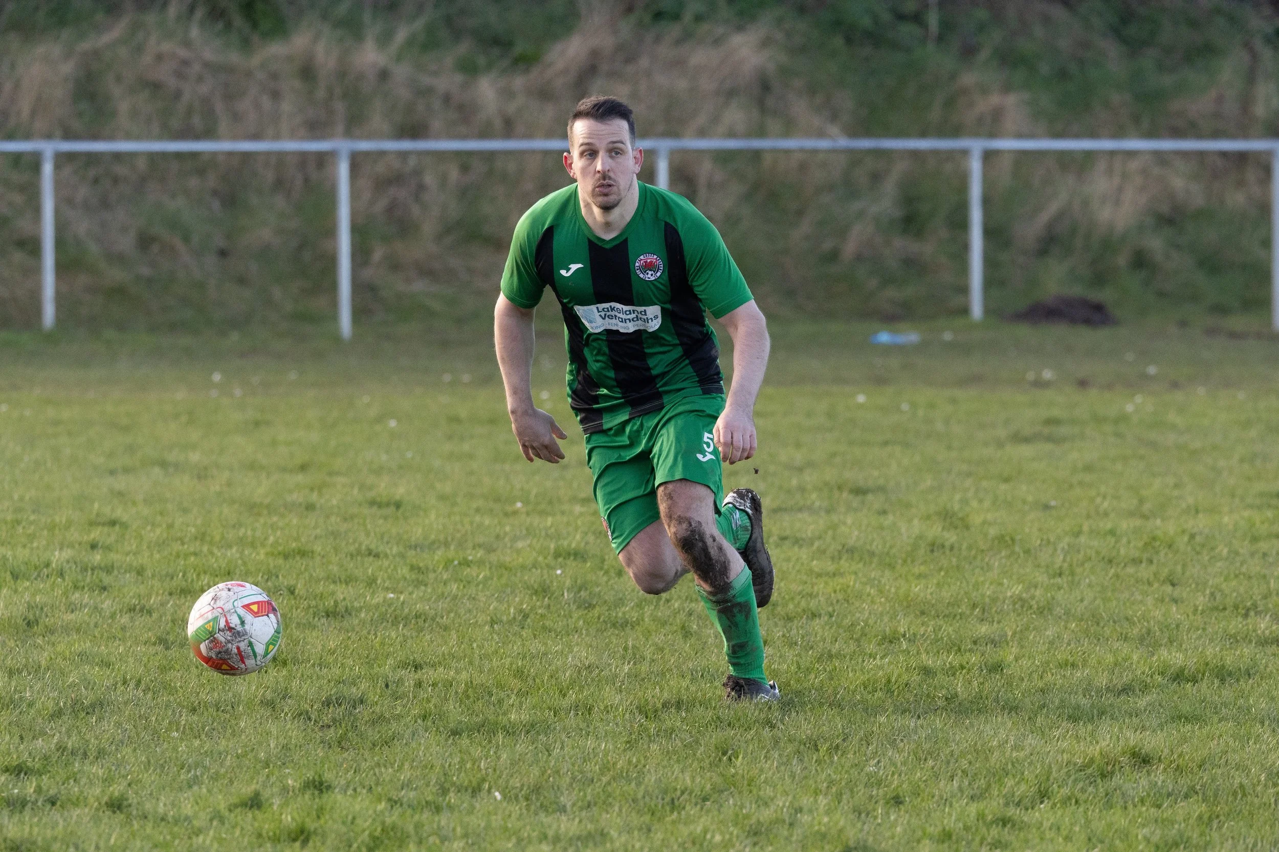 A man playing soccer on a grassy field, wearing a green and black uniform with the number 5, running towards the ball, which is on the ground.