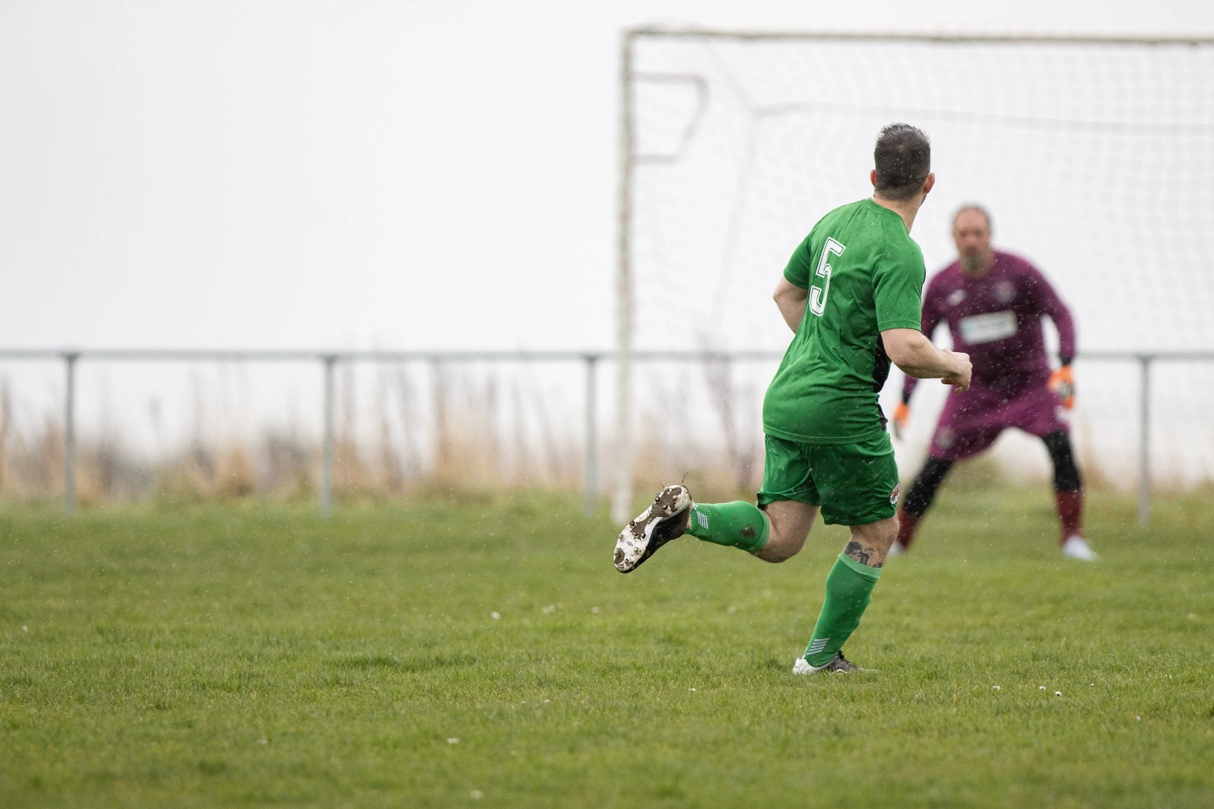 Soccer player in green jersey with number 5 running towards goal as goalkeeper in purple uniform stands in front of goalpost