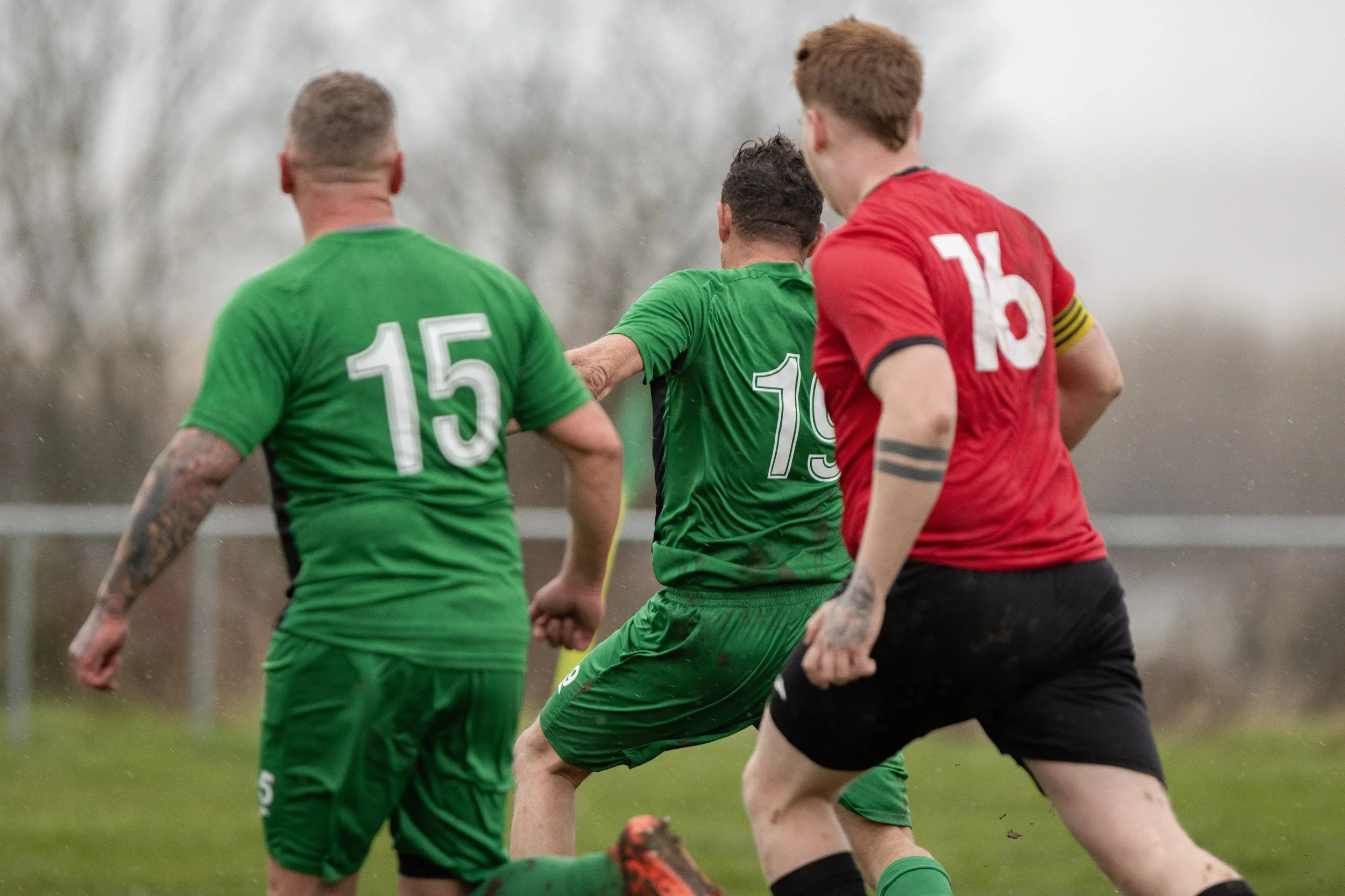 Soccer match in progress with three players in focus on the field, two in green jerseys and one in a red jersey, during a cloudy day.
