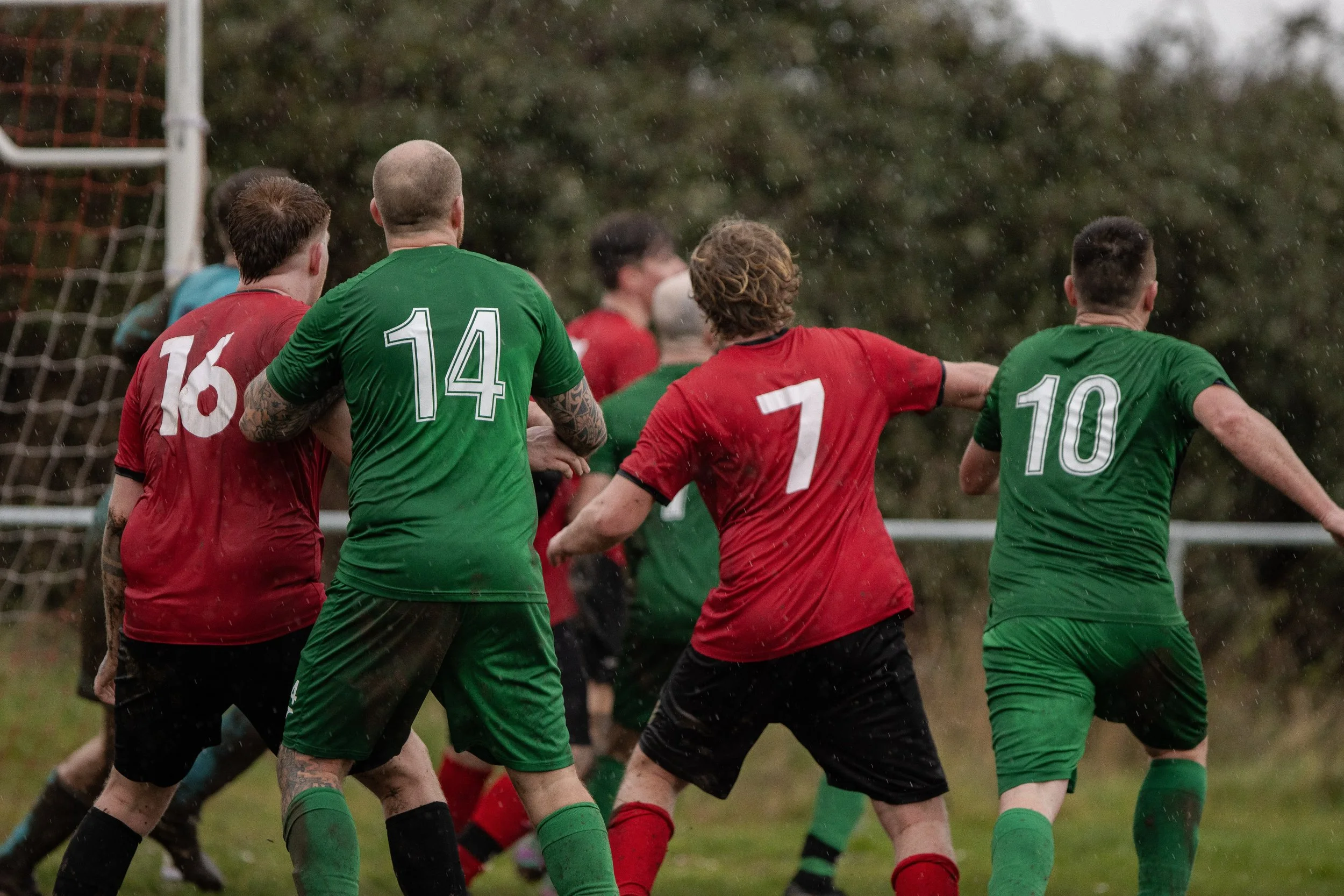 Soccer players in red and green jerseys competing in the rain during a match, near the goal.