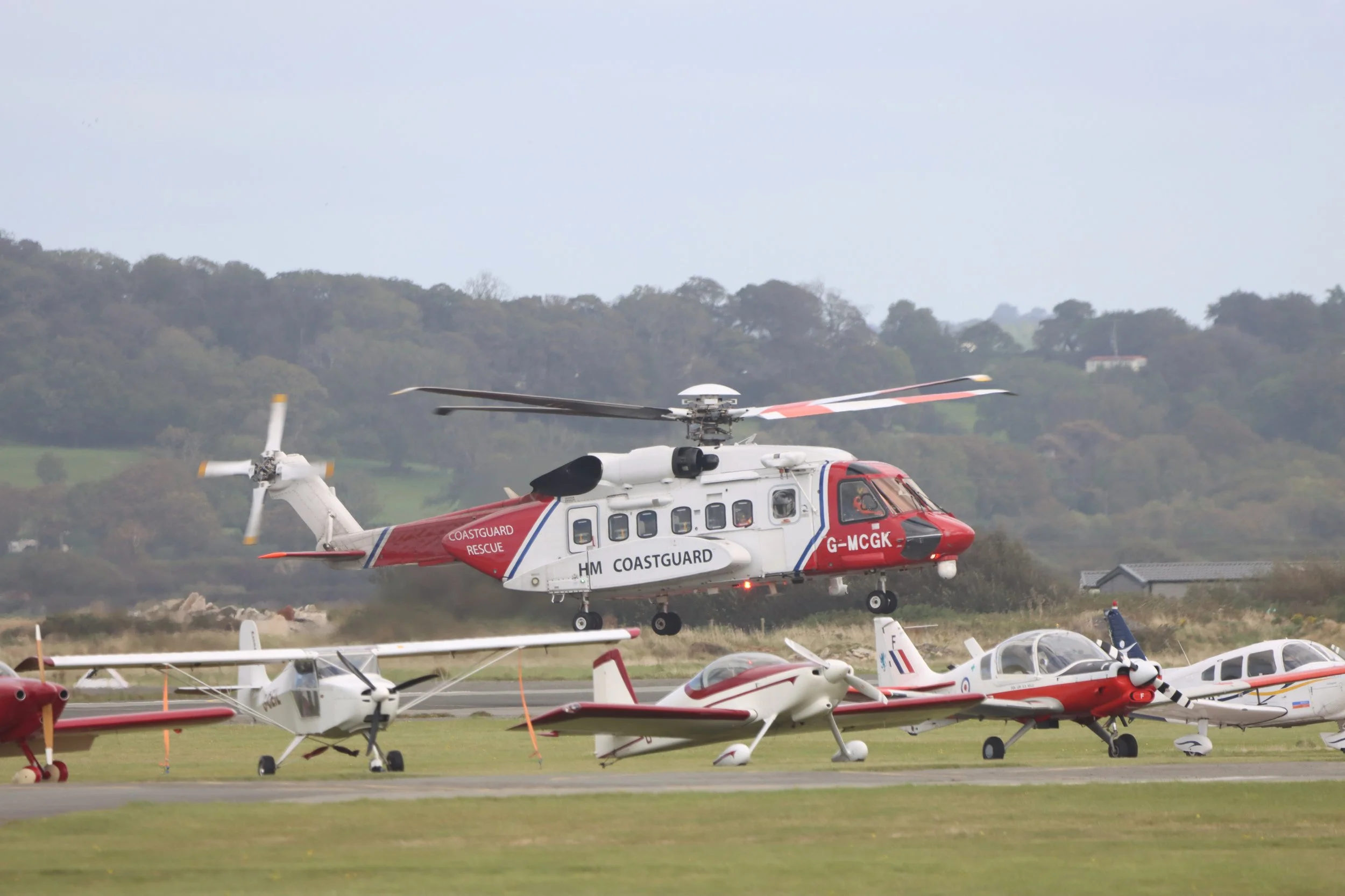 A helicopter with Coast Guard RESCUE markings flying over small fixed-wing aircraft parked on a runway with a grassy field and trees in the background.