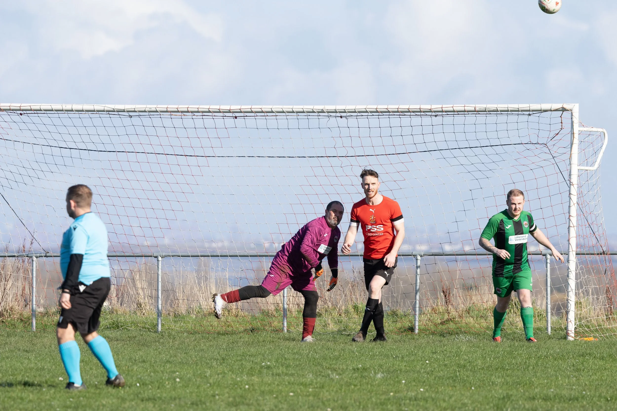 Soccer players on the field during a game, near the goal, with a goalkeeper in a purple uniform and two outfield players in red and green jerseys, on a grassy field under a cloudy sky.