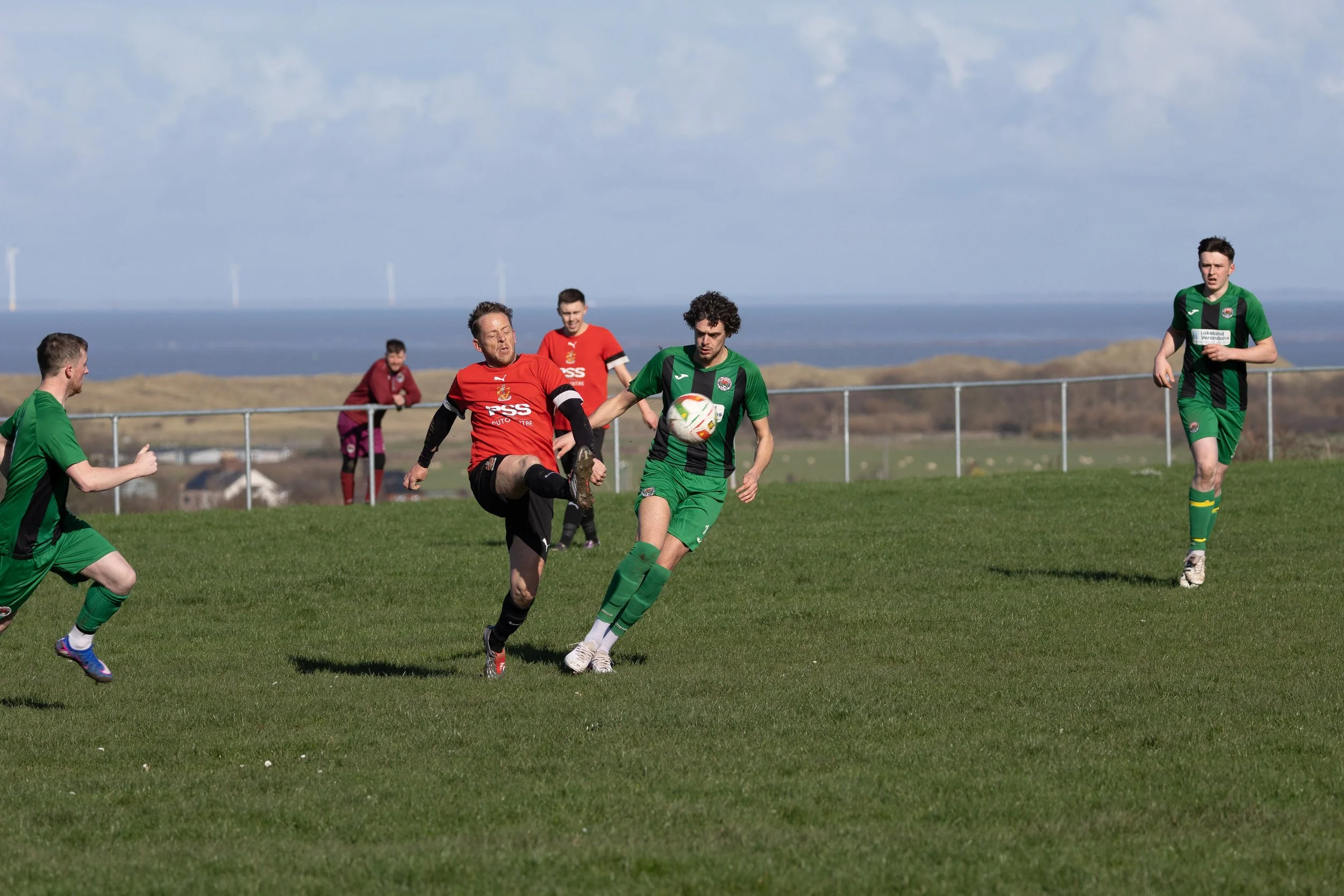 Soccer game with players in green and red jerseys playing on a grass field near the coast.