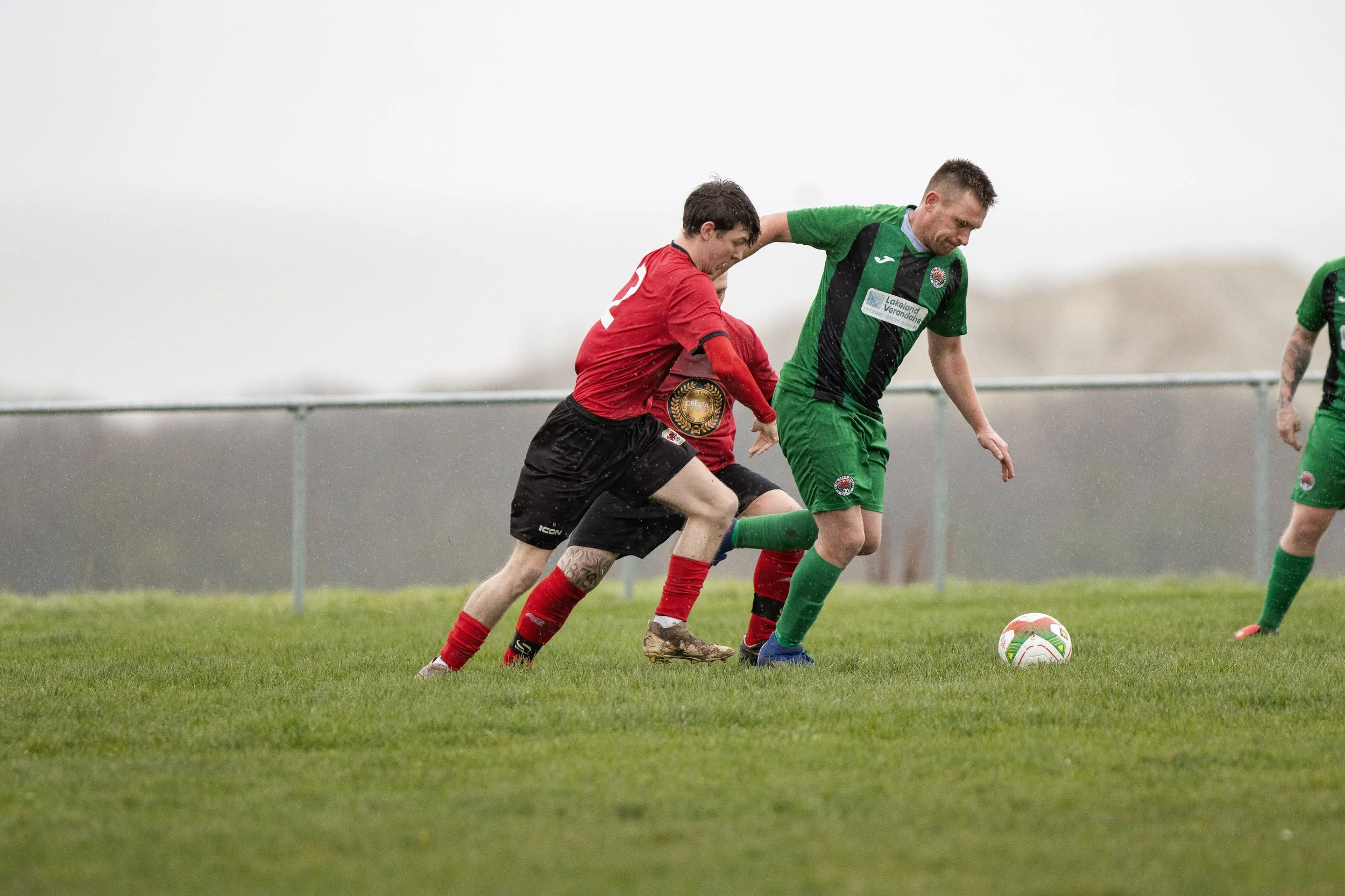 Two soccer players competing for the ball on a grassy field, one in red and black, the other in green and black, in rainy weather.
