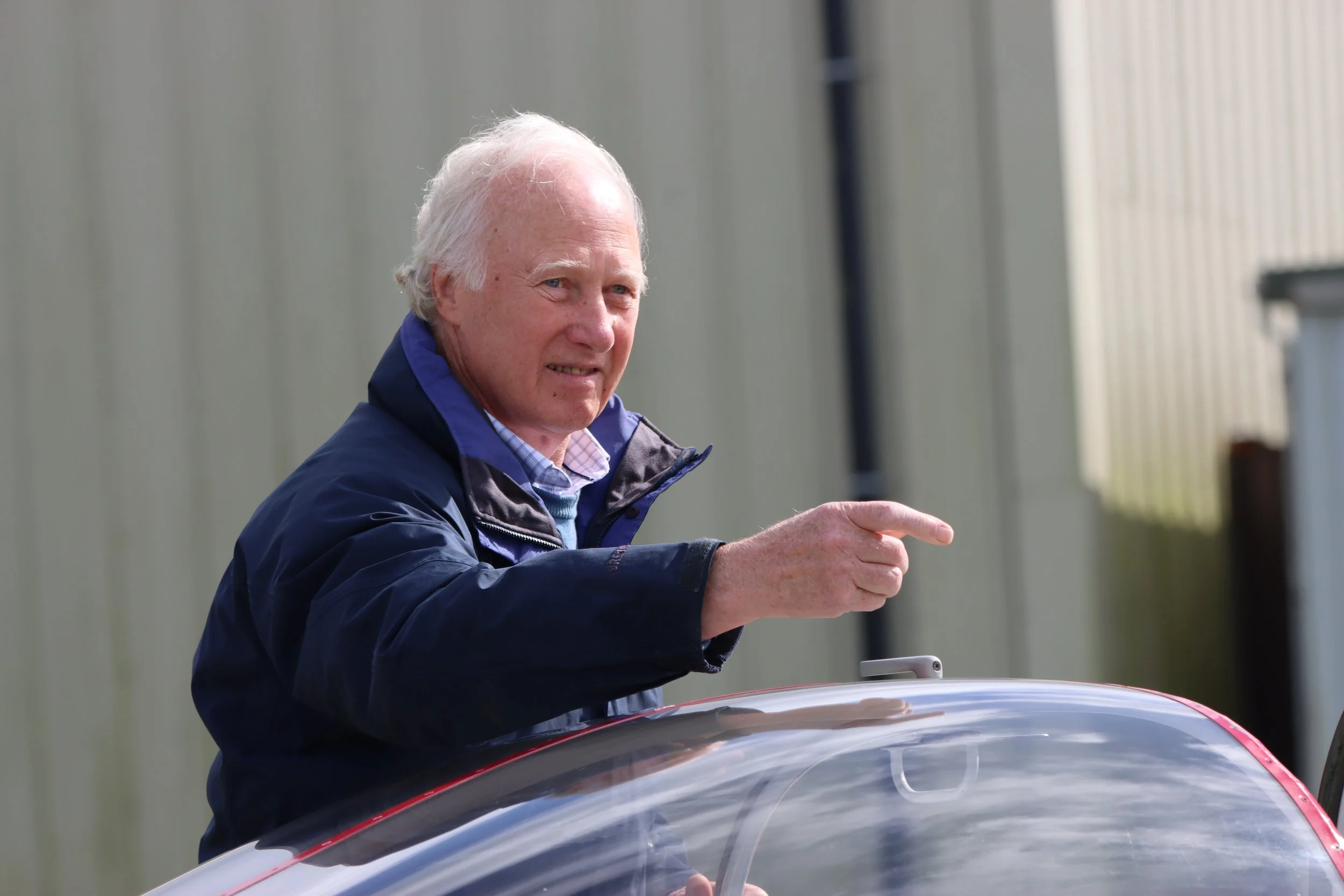An elderly man with white hair, wearing a blue jacket, is pointing and smiling while standing next to a small aircraft or glider with a clear canopy.