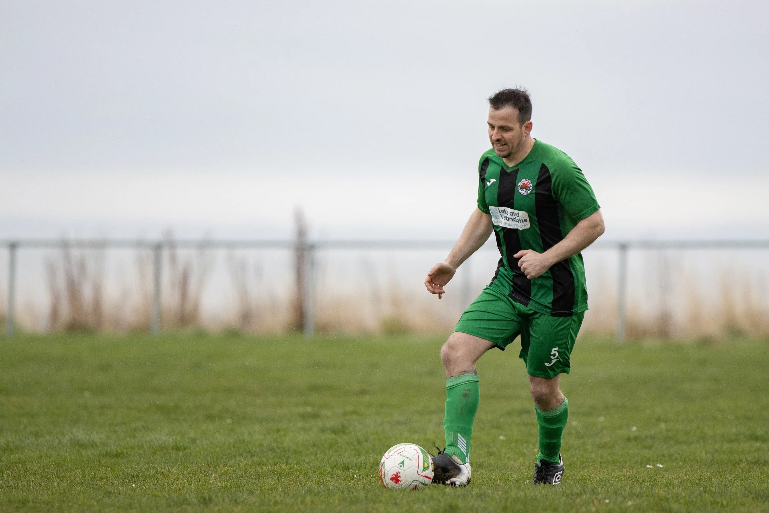 A male soccer player in a green and black uniform, wearing the number 5, is on a grassy field preparing to kick a soccer ball.