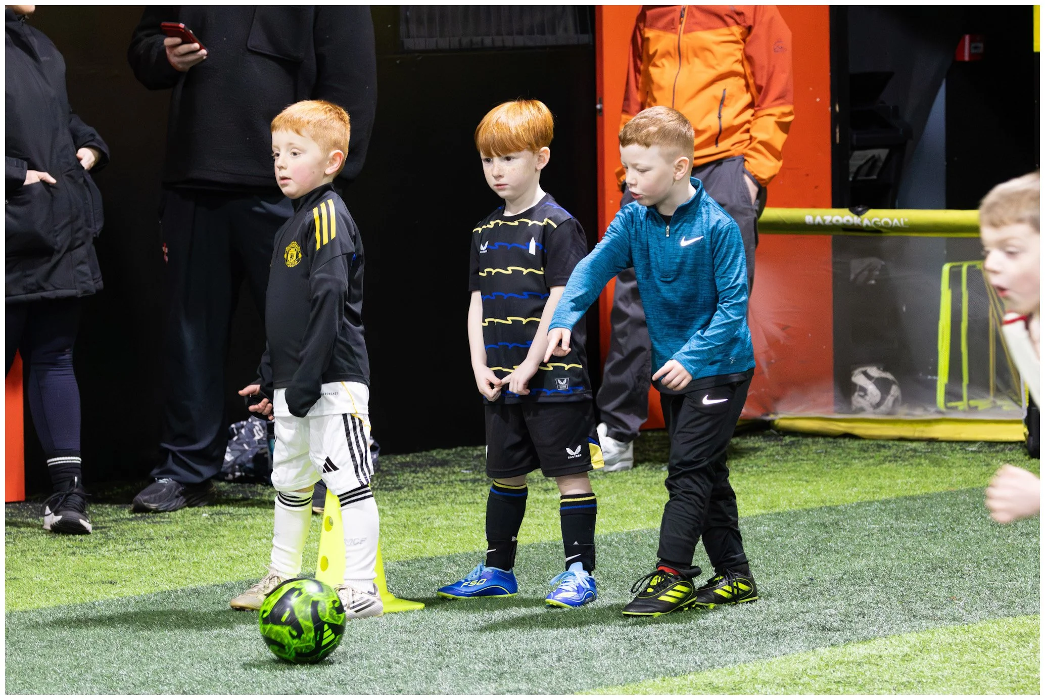 Three young boys standing on an indoor soccer field, with one boy preparing to kick a neon green and black soccer ball, while other children and adults observe.