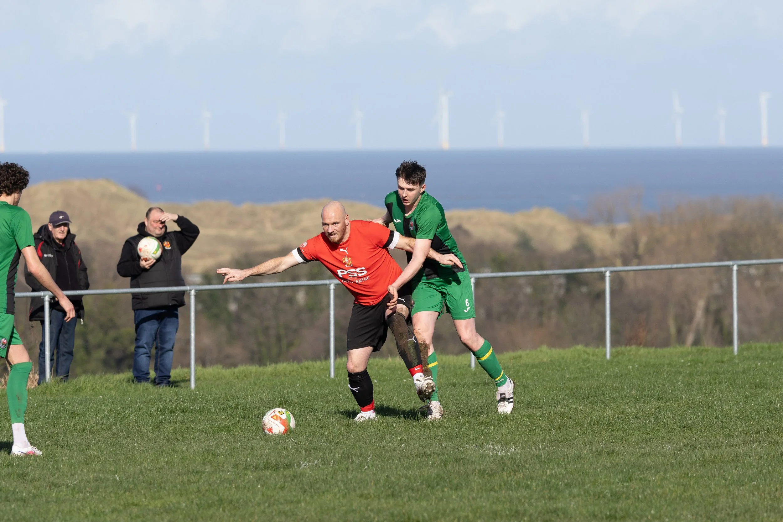 Two soccer players compete for the ball during a match on a grassy field, with a scenic background of hills and wind turbines in the distance.