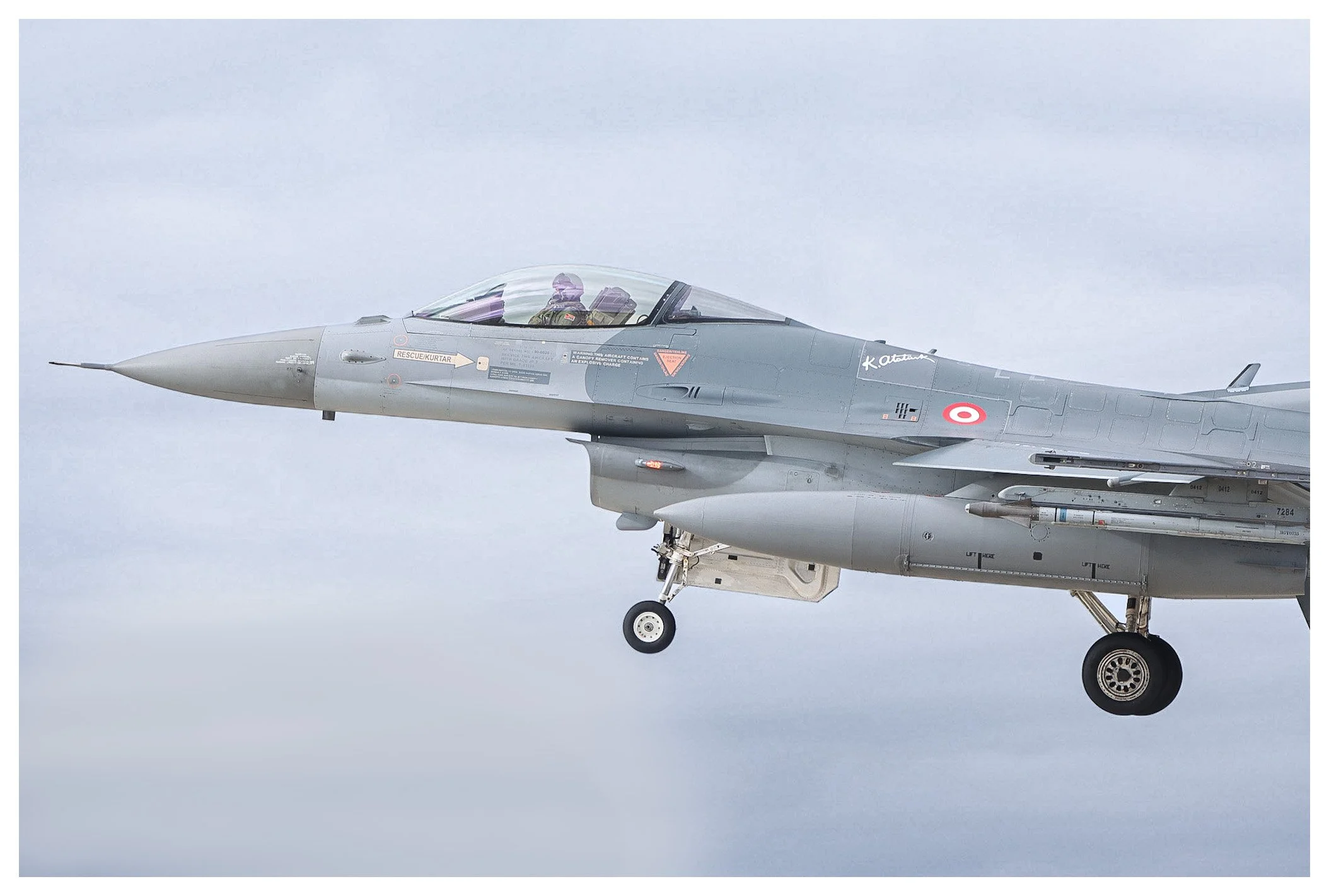 A military fighter jet aircraft flying in the sky with gray clouds, seen from the side with the landing gear down.