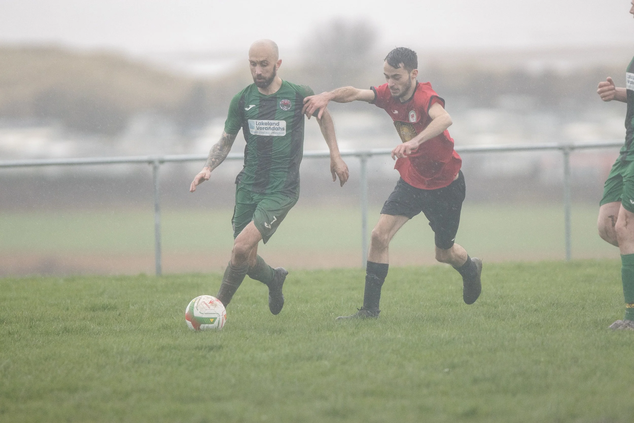 Two soccer players competing for ball on rainy field with foggy background.