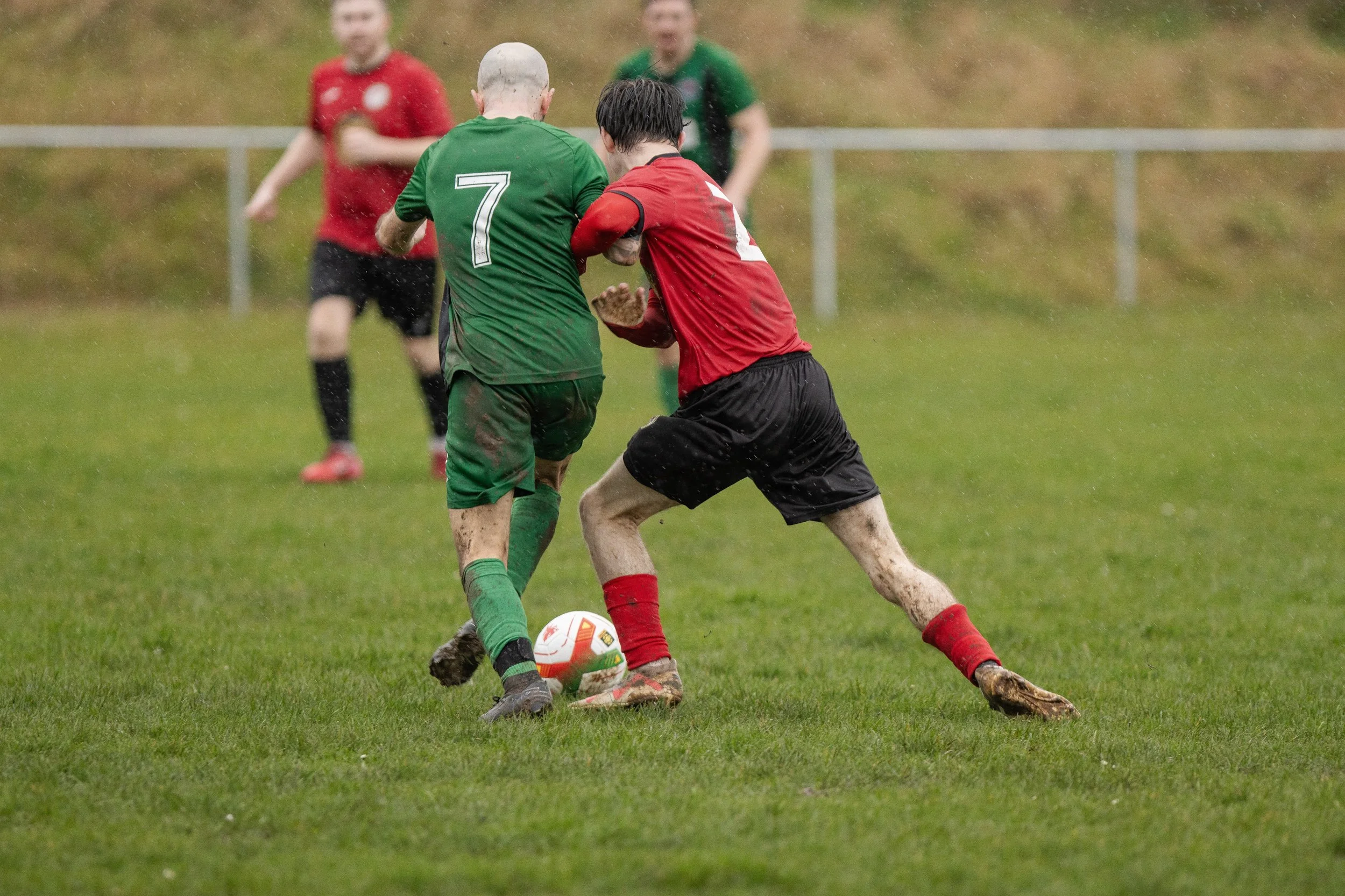 Two soccer players, one in a green jersey with the number 7 and the other in a red jersey, are fighting for the ball on a muddy field in the rain, with other players and a grassy hill in the background.