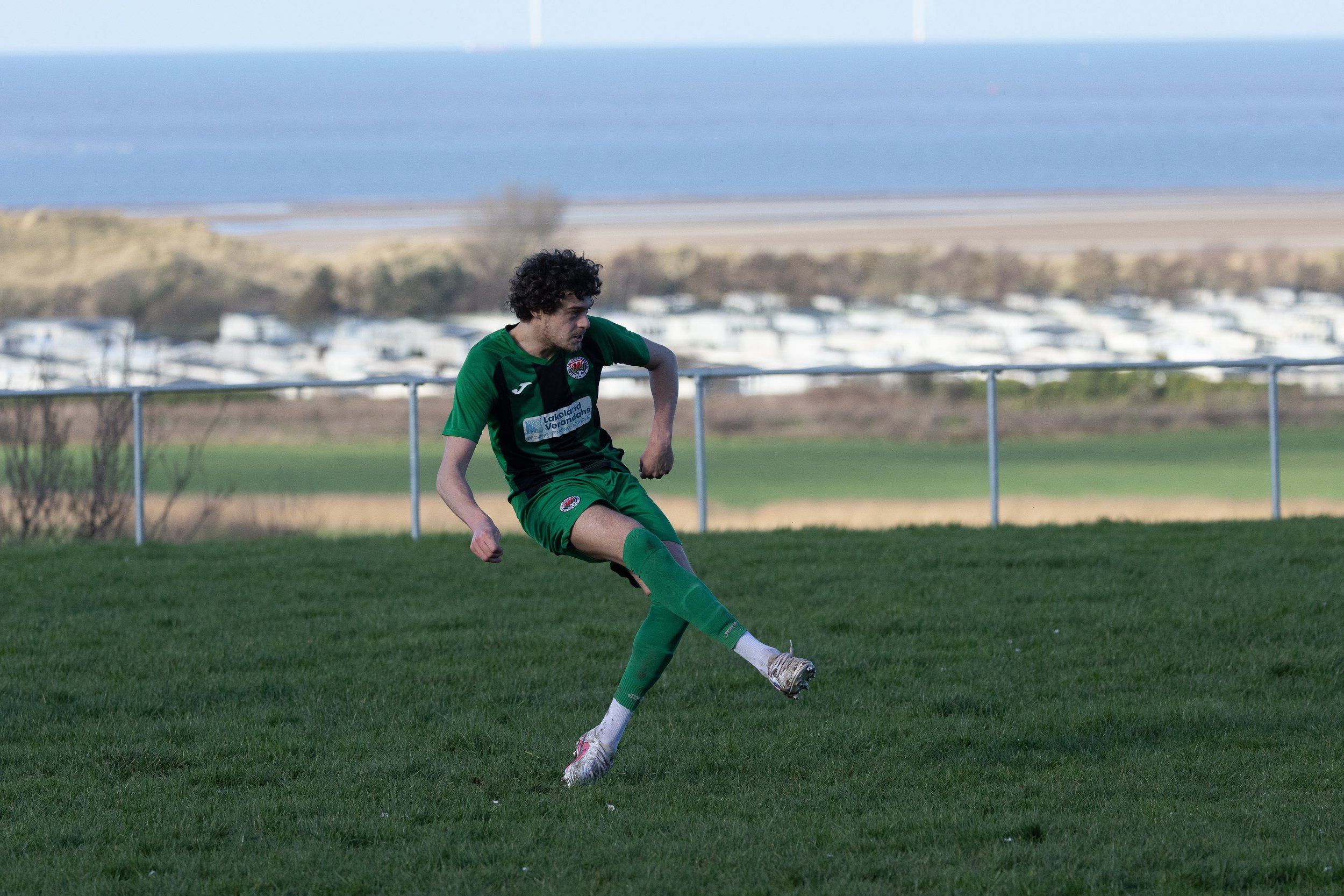 A soccer player in a green uniform kicking a ball on a grassy field with a coastal background.