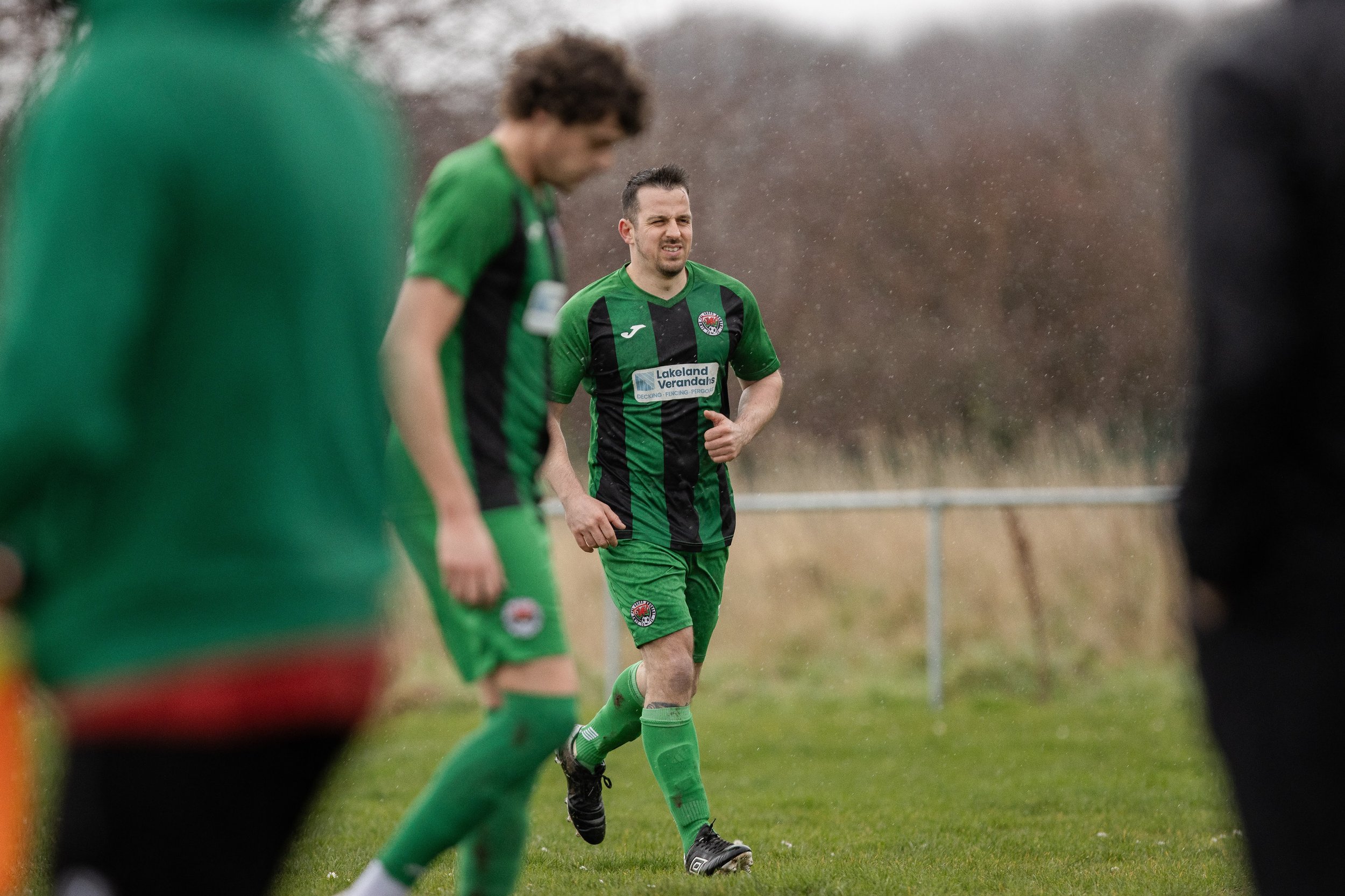 A group of soccer players in green and black uniforms on a grassy field, with one player in the center jogging, rain falling, and a blurred background of trees and a fence.