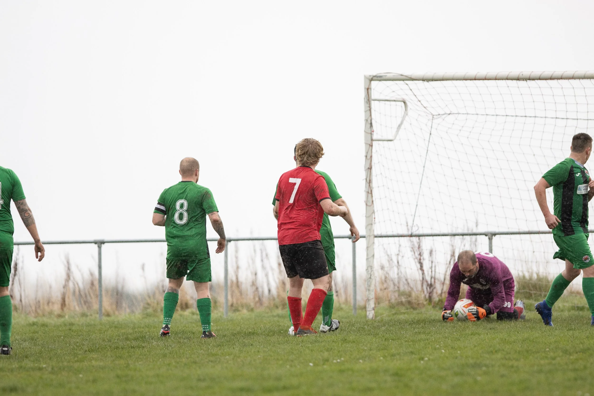 A soccer game with players in green and red jerseys near the goal, where the goalkeeper in purple is holding the ball after a save on a cloudy day.