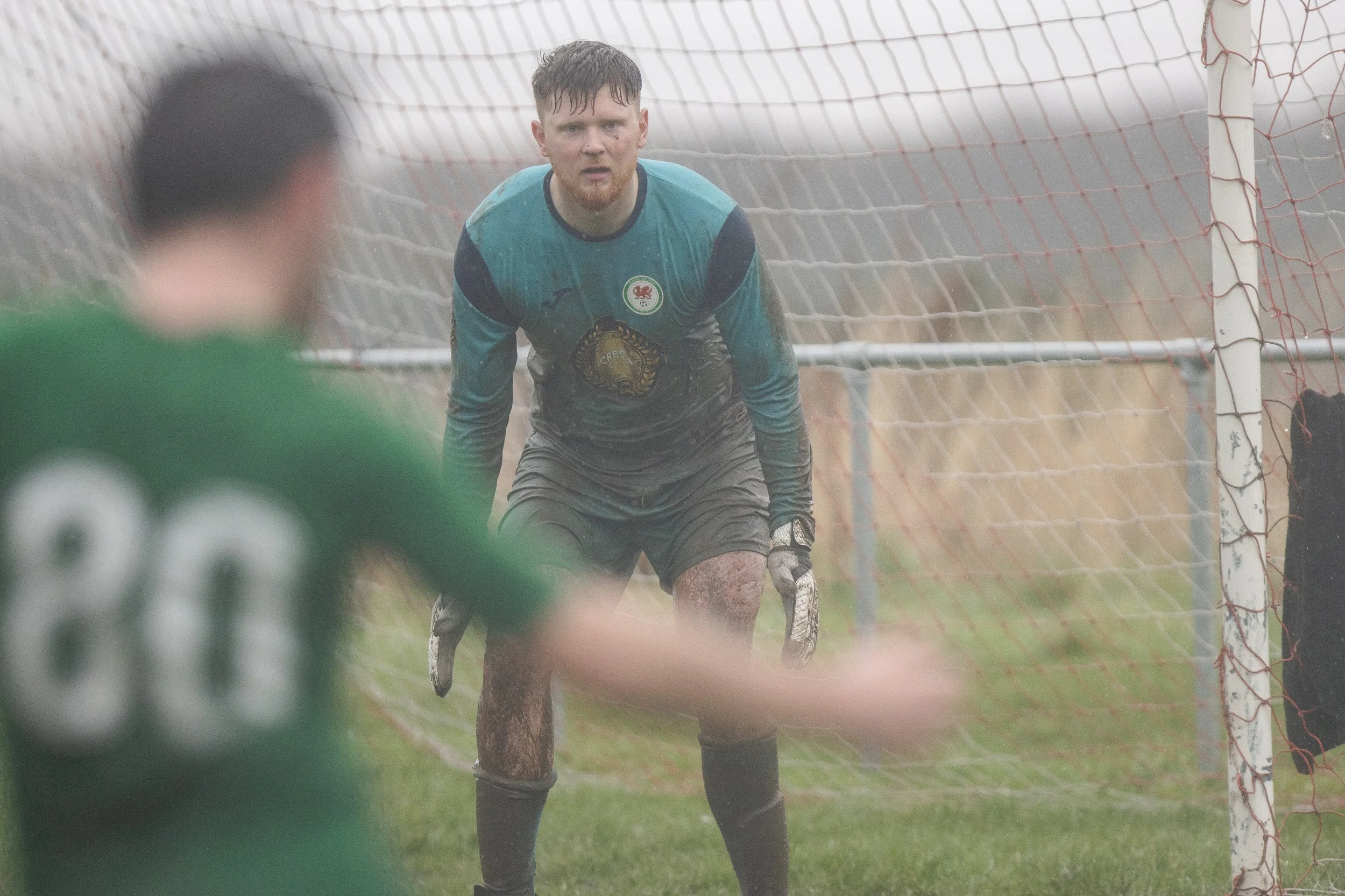 A soccer goalkeeper in a blue and black kit is on the field in front of the goal, looking focused and ready to make a play, with rain falling around them.
