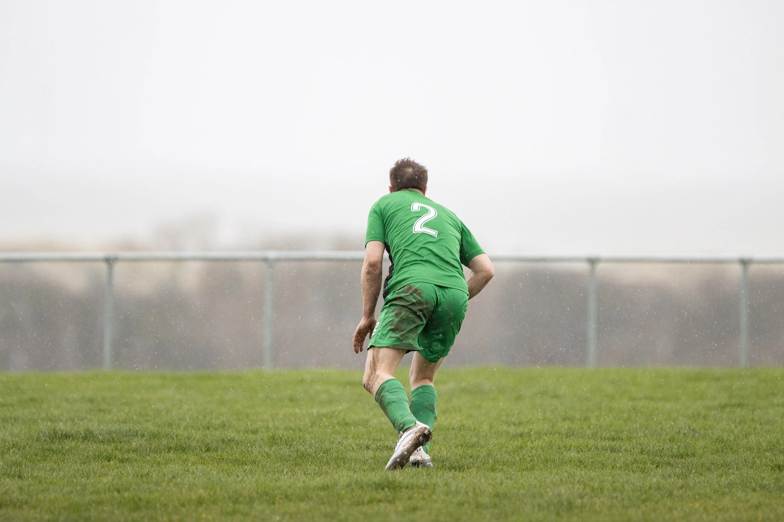 A soccer player dressed in green uniform with the number 2 on his back, running on a grassy field during a game on a rainy day.