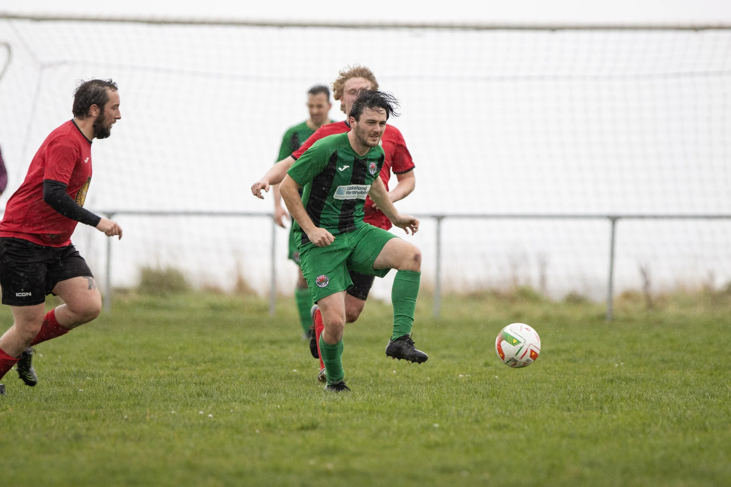 Soccer players playing on a grassy field during rain, with one player in a green uniform controlling the ball while others in red are nearby.