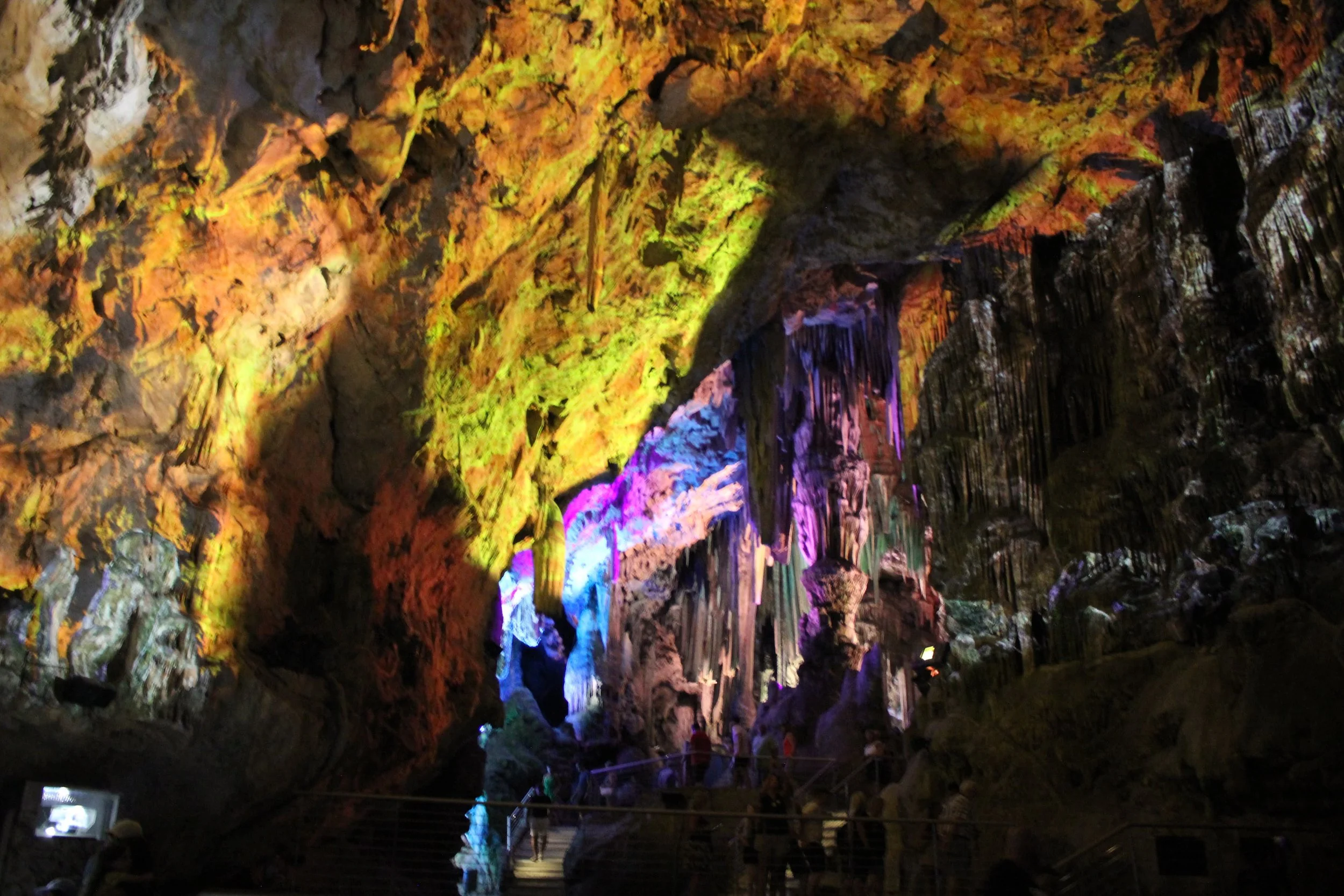 Interior view of a large cave with colorful stalactites and stalagmites illuminated by multicolored lights, with visitors walking on a pathway inside.