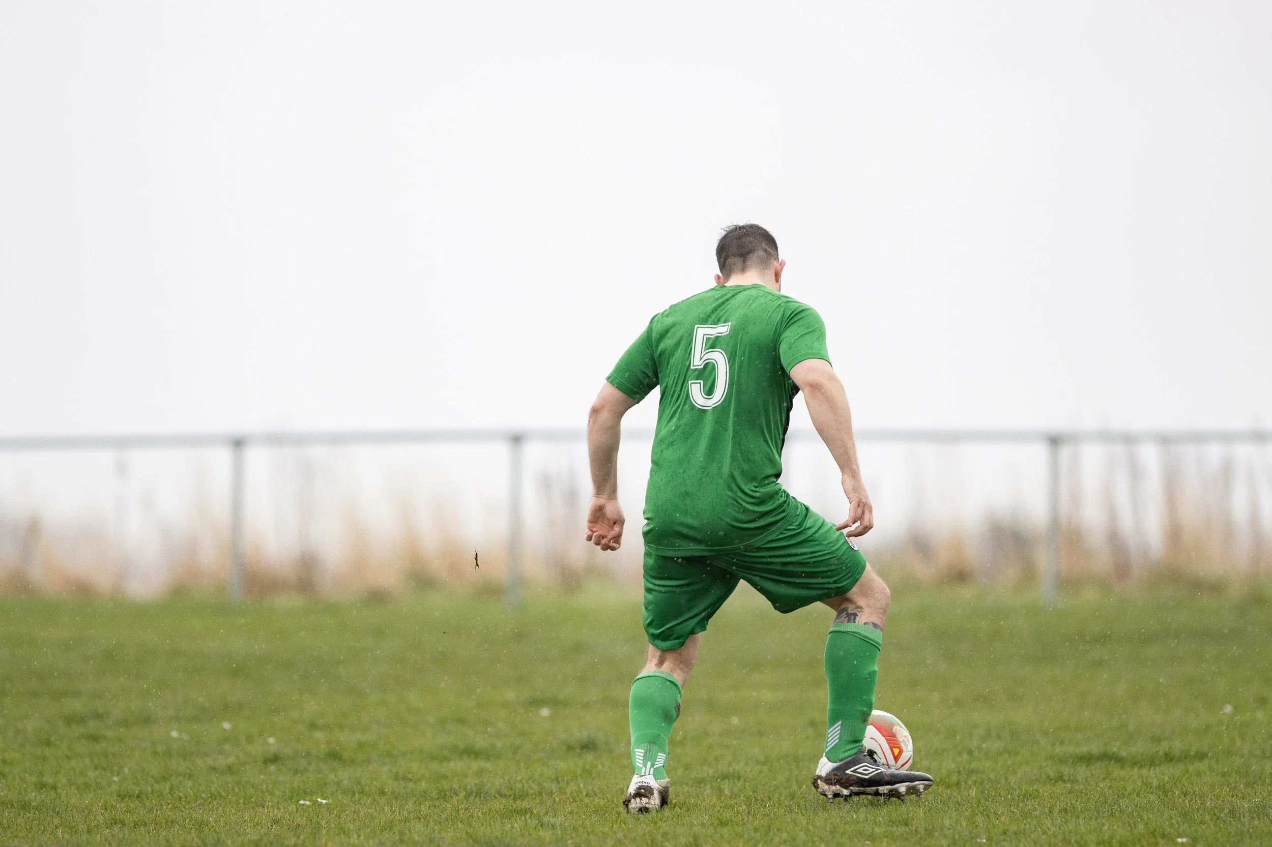 A soccer player in a green uniform with the number 5 on his back, standing on a grass field and preparing to kick a soccer ball.