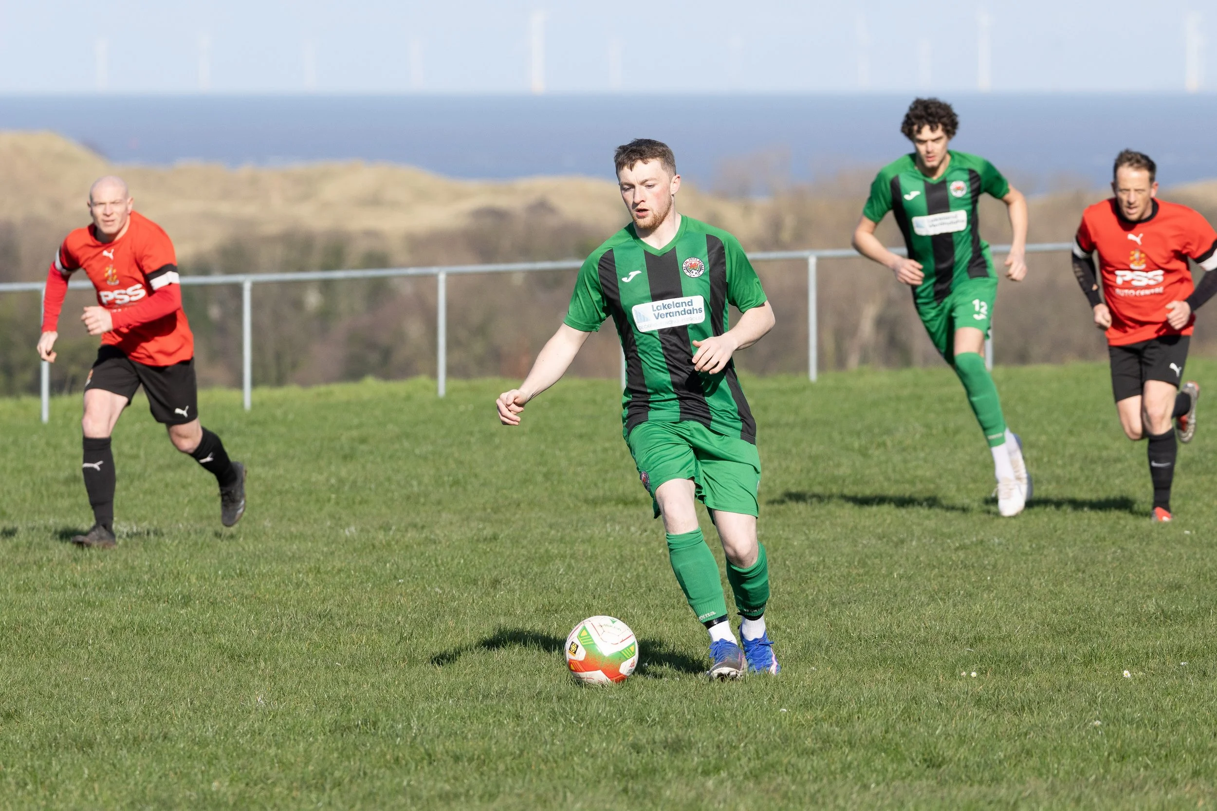 Soccer players on a grassy field, with five individuals in action. One player in a green and black jersey is in the foreground about to kick the ball, while others in red and black jerseys are running behind him, with hills and wind turbines in the b
