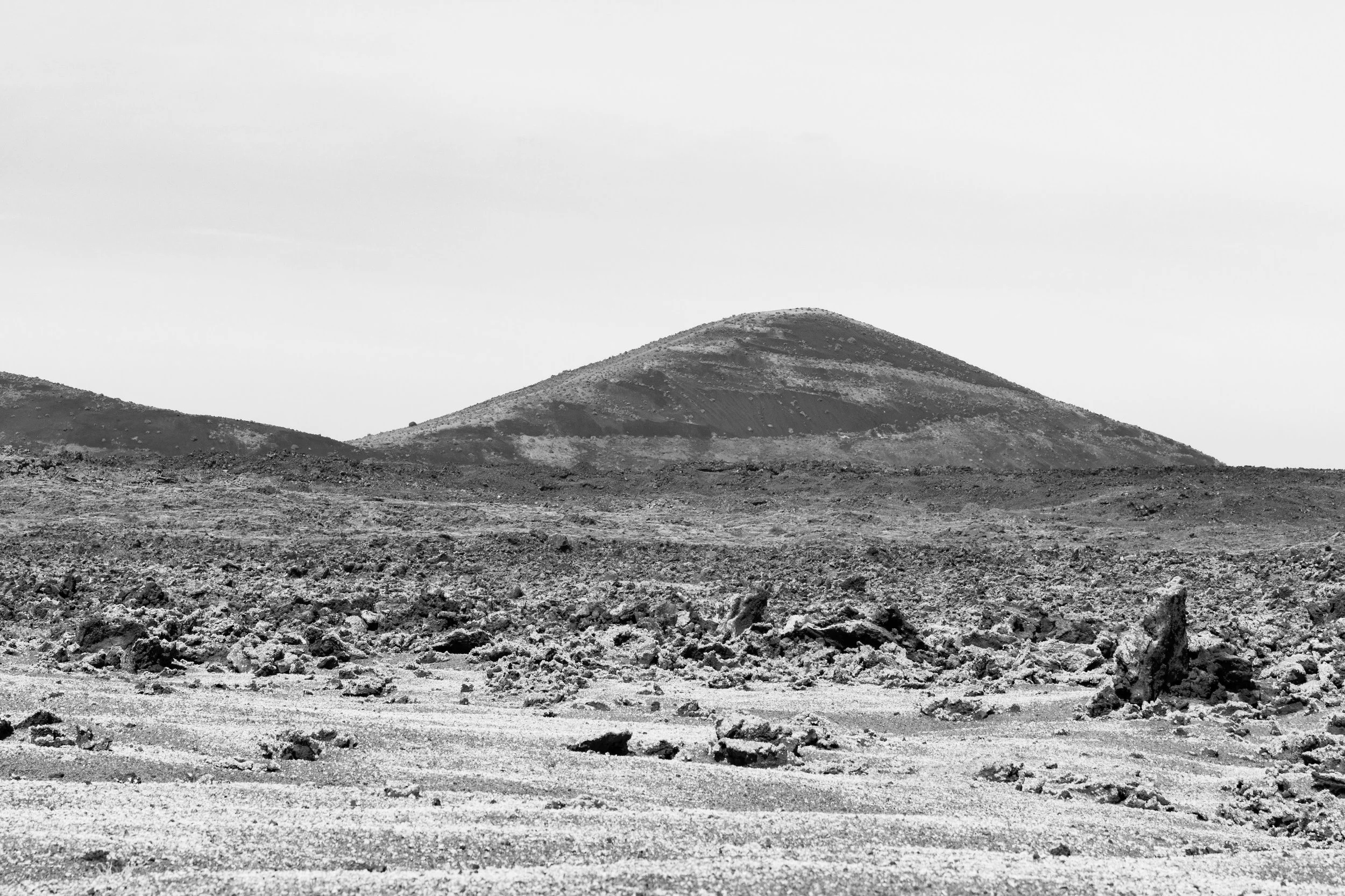 Black and white photo of a barren, rocky landscape with a small mountain in the distance under a cloudy sky.