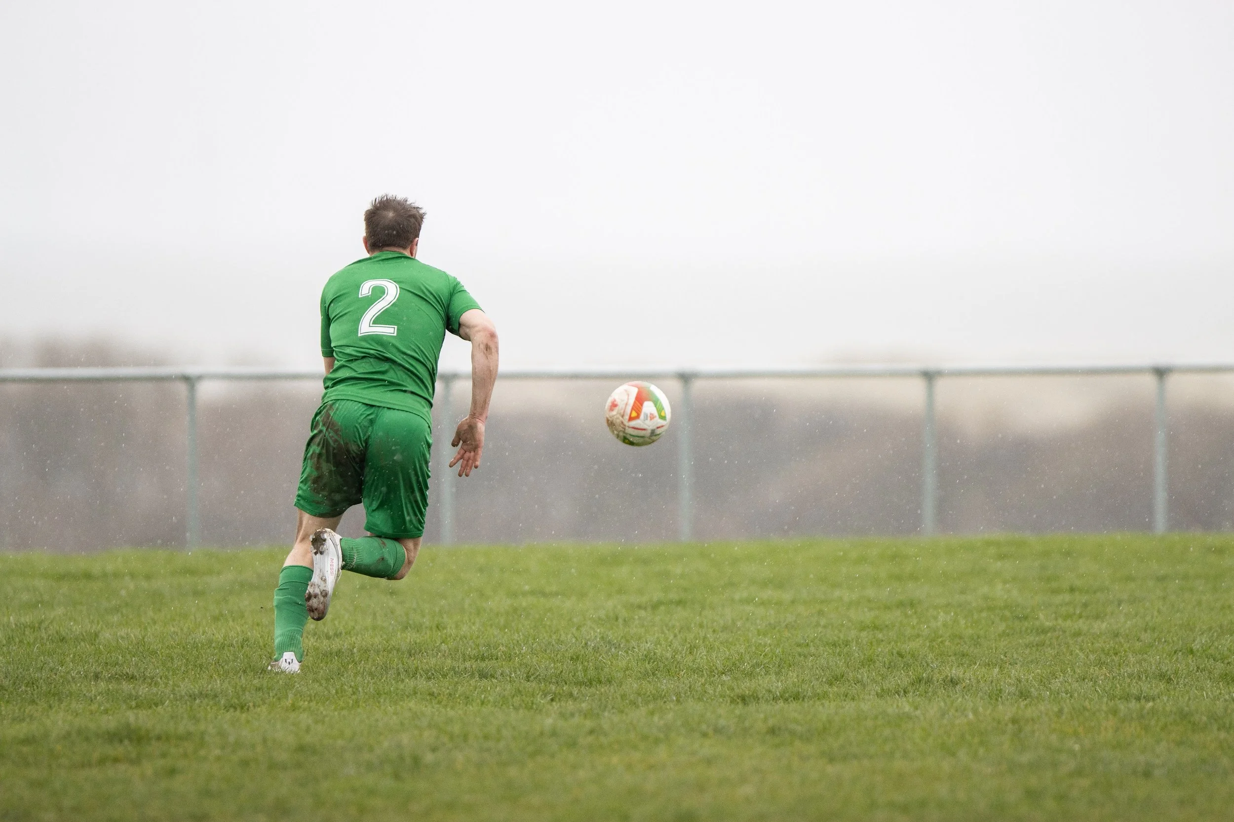 A soccer player in a green uniform running on a field, with the number 2 on his back, as the ball is in mid-air in front of him.