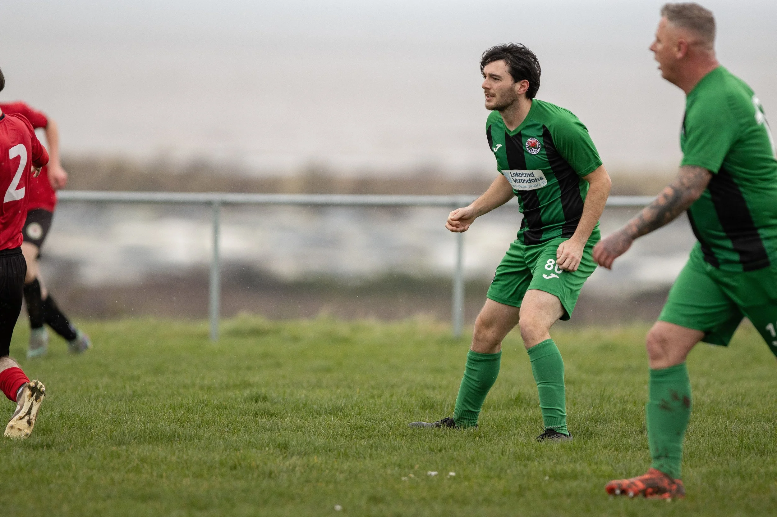 Soccer players on the field, with two players in green jerseys and one in a red jersey, playing on grassy field with cloudy sky background.