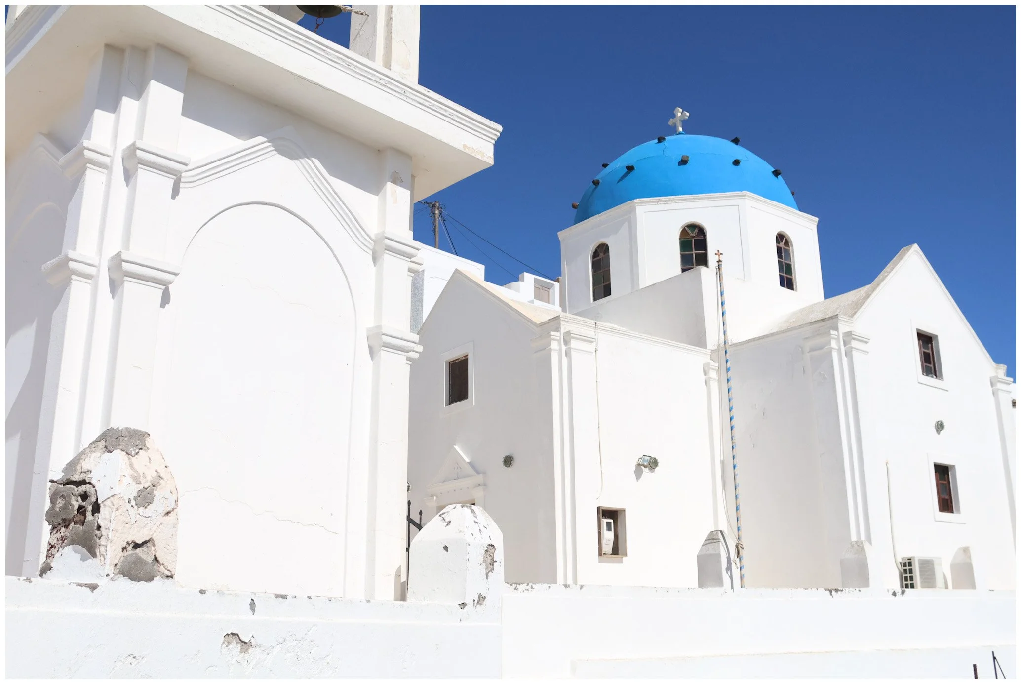 White Greek church with blue dome and cross, against a clear blue sky.