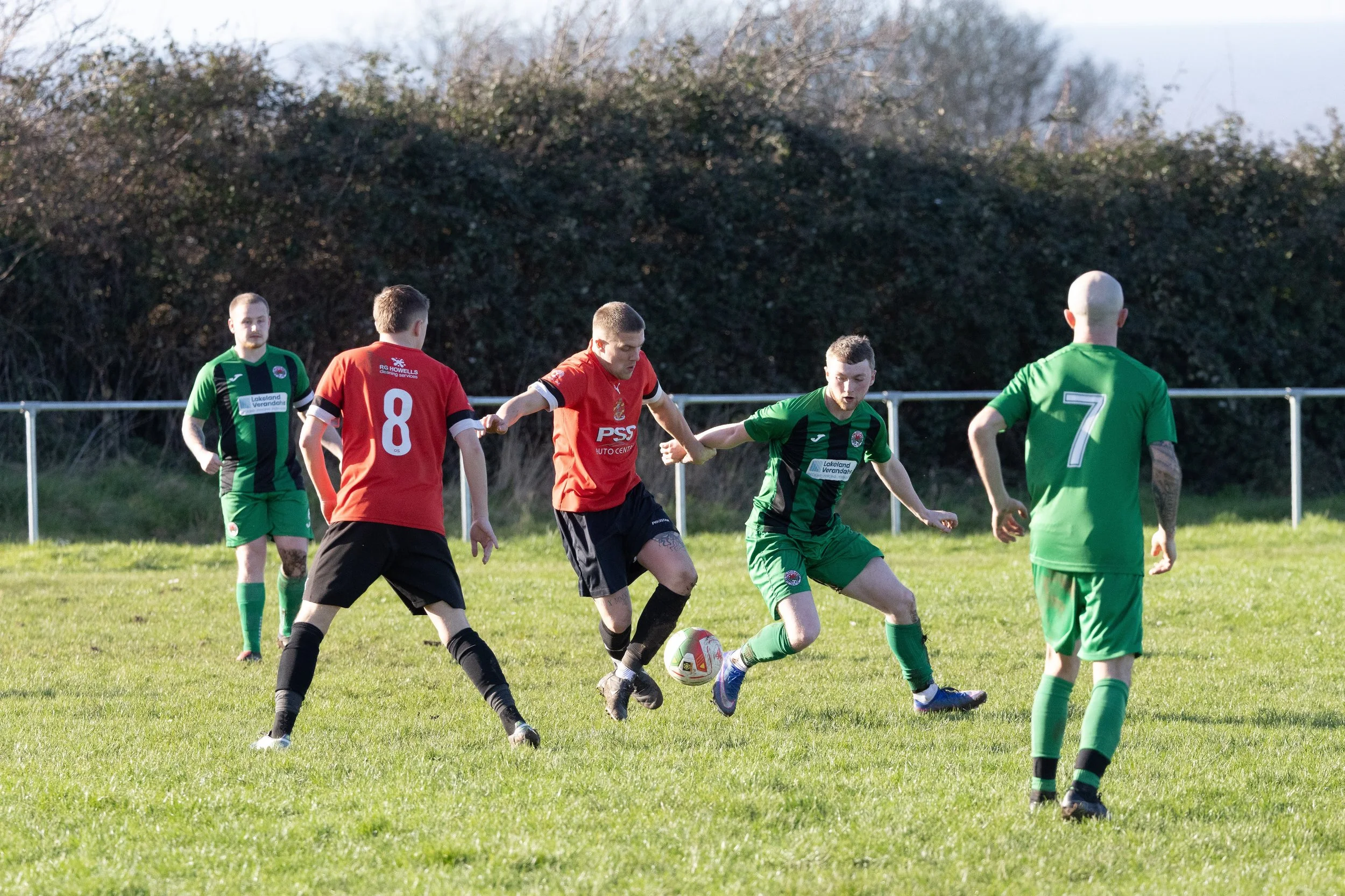 Soccer players in green and red jerseys competing for the ball on a grassy field.
