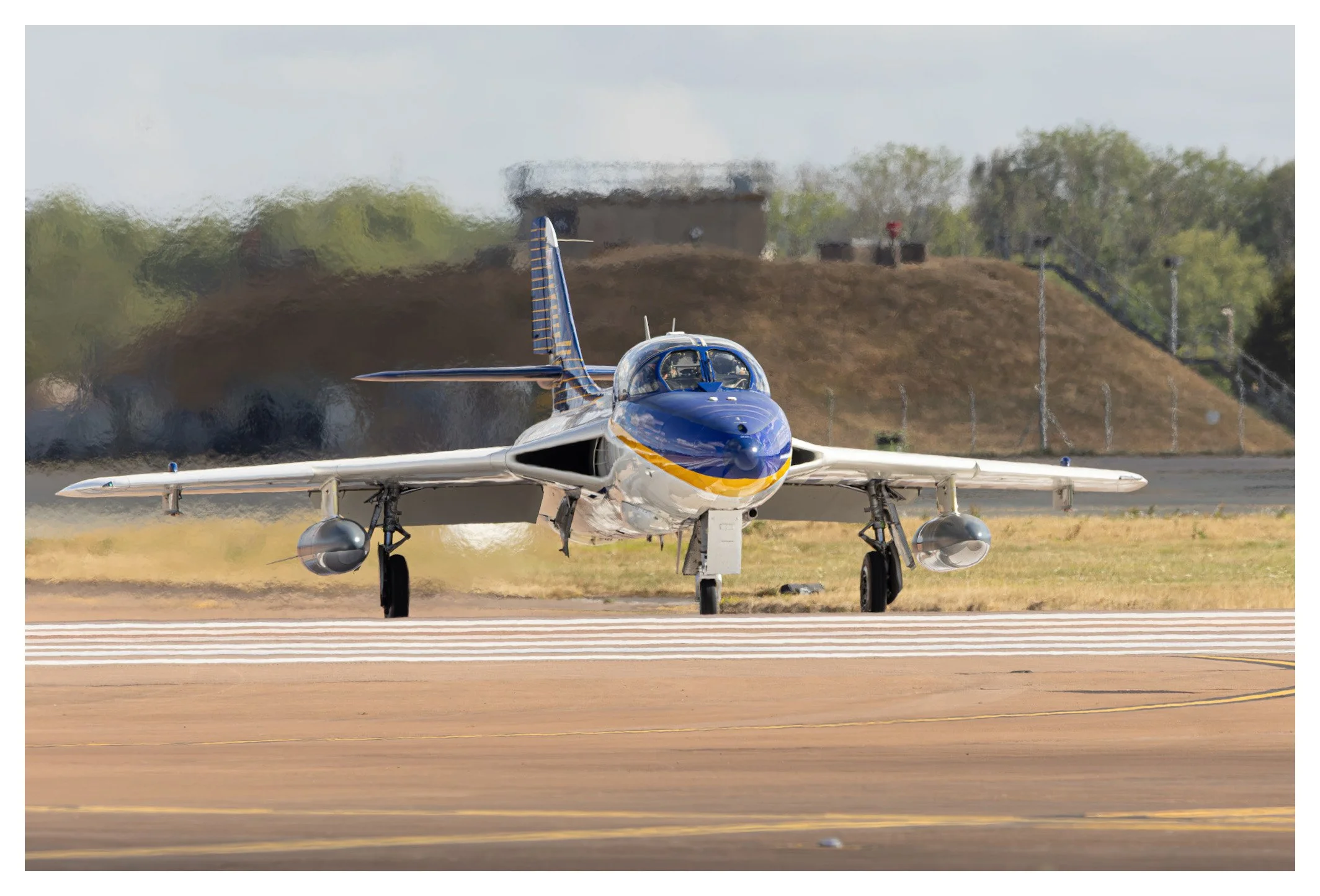 A military jet aircraft on the runway at an airfield, ready for takeoff or landing, with a hill and guardrail in the background.