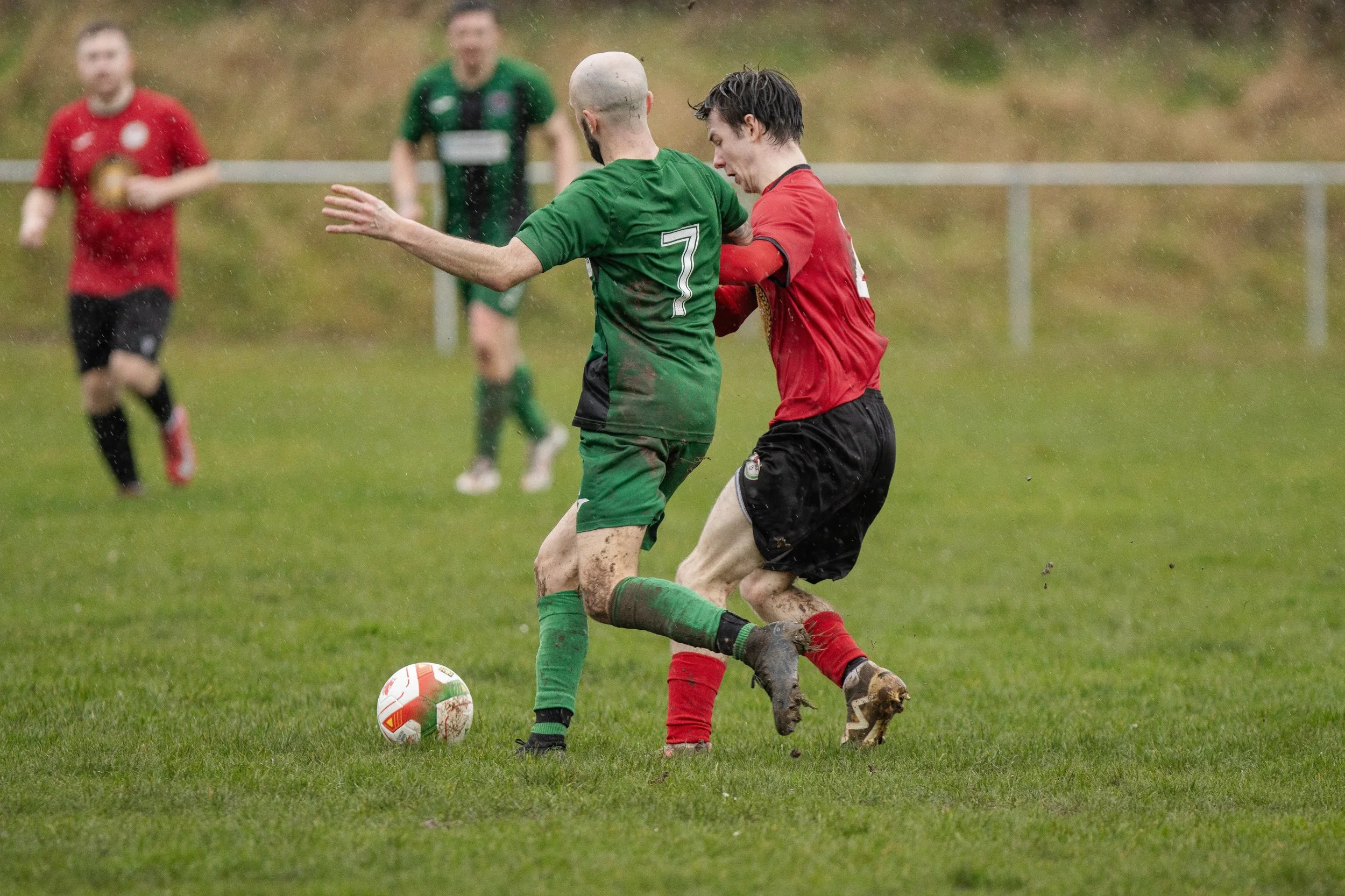 Two soccer players battling for the ball on a muddy field with wet weather, with additional players blurred in the background.