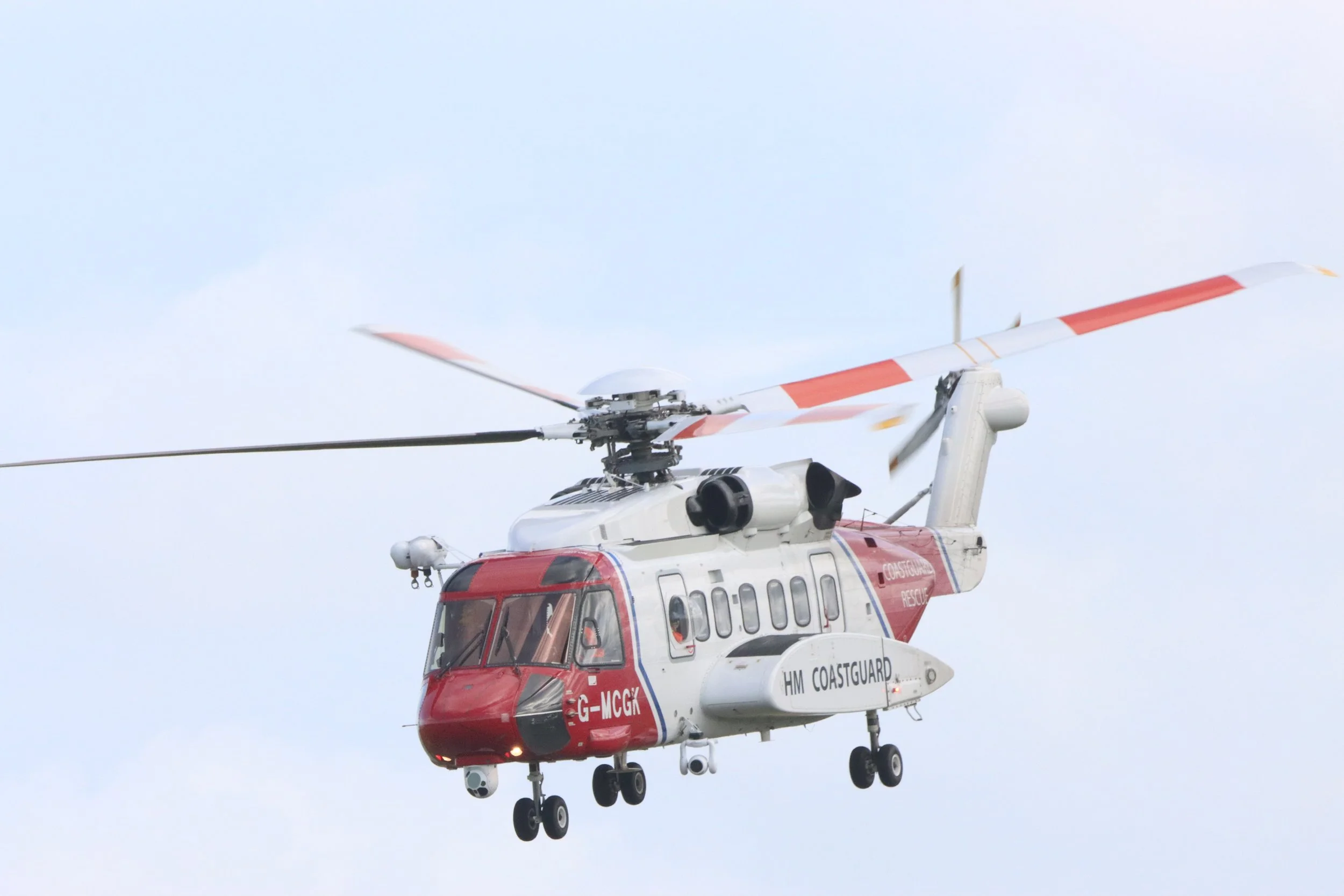 A helicopter in flight with a white and red color scheme, labeled HM Coastguard, flying against a cloudy sky.