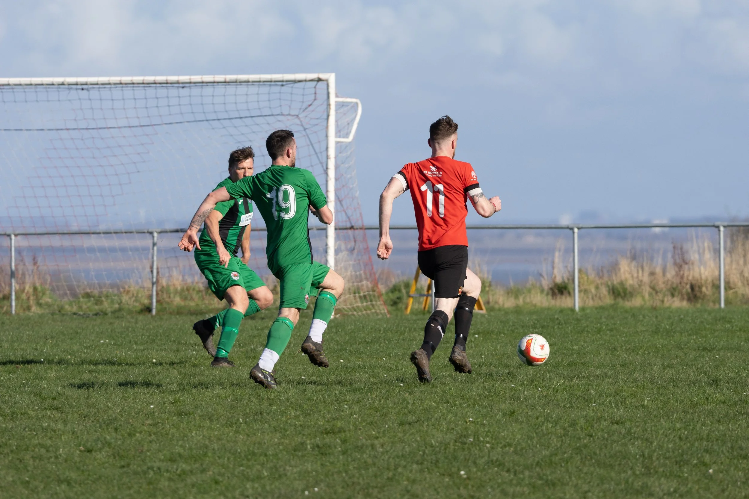 Three soccer players, two in green jerseys and one in a red jersey, running towards the goal with a soccer ball on a green field under a partly cloudy sky.