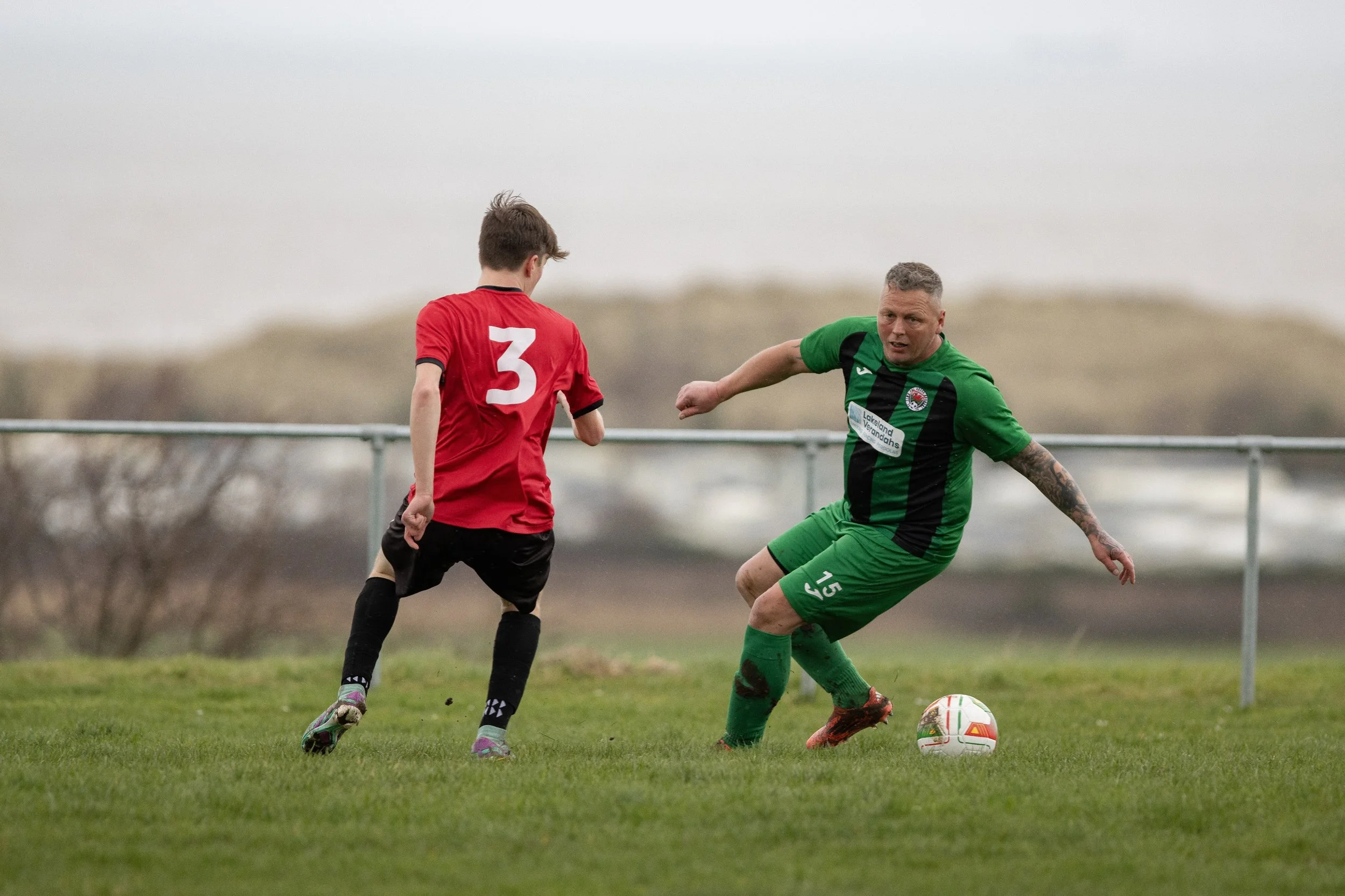 A soccer player in a green and black uniform is kicking a soccer ball while approaching a young player in a red jersey with the number 3 during a game on a grass field with a cloudy sky.