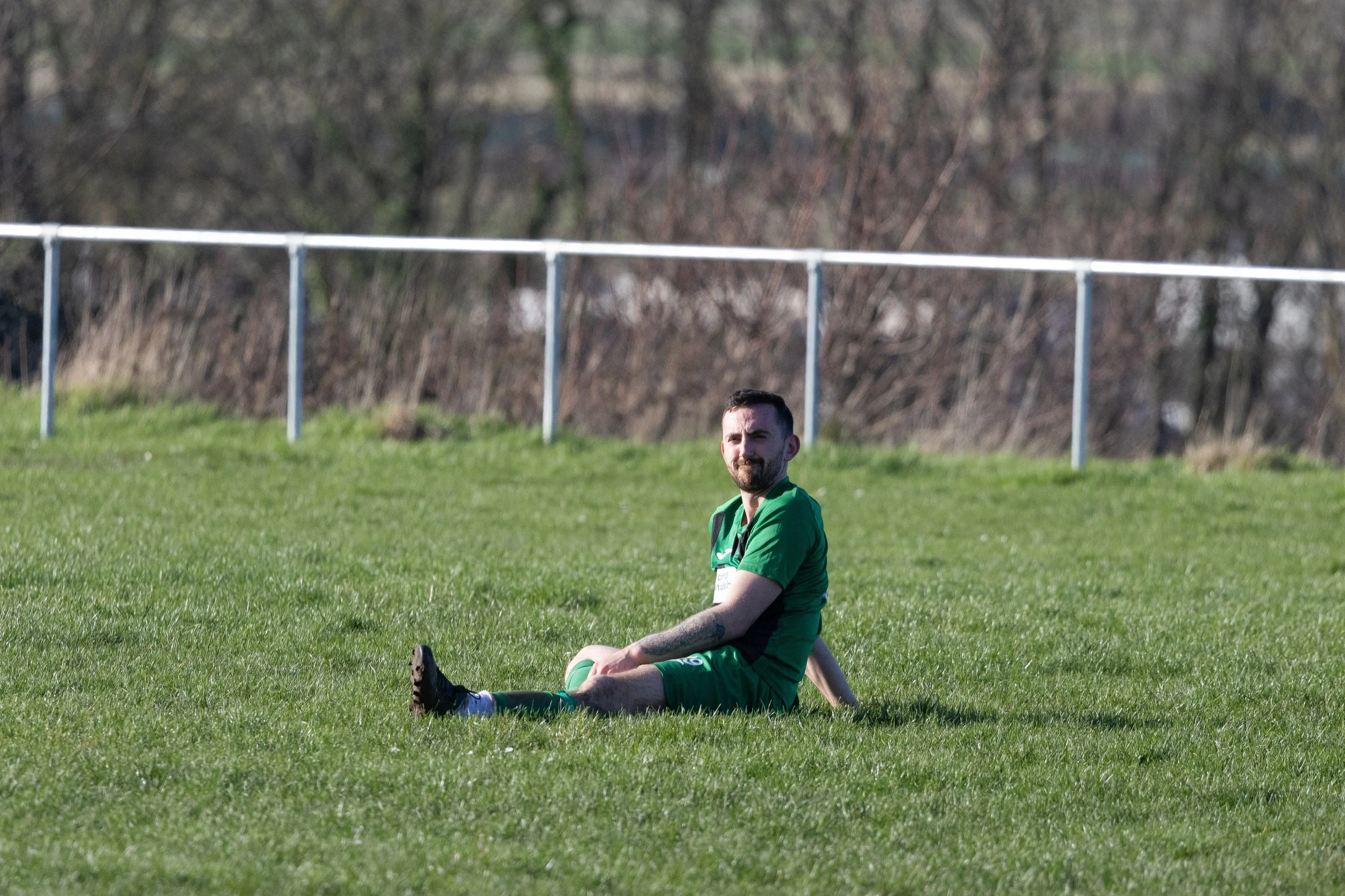 A man with a beard in a green soccer uniform sitting on the grass field during daytime, with a white railing and trees in the background.