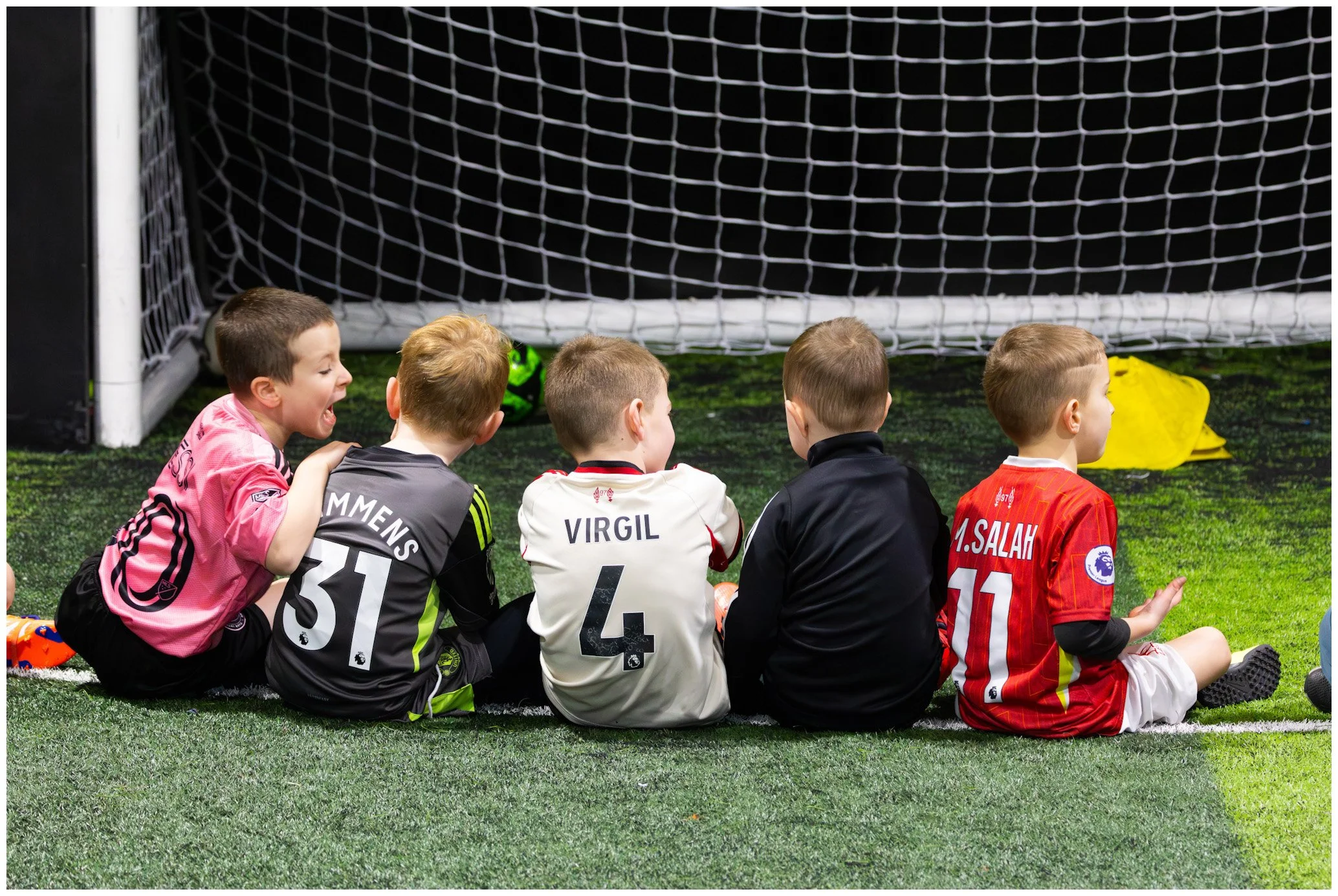 Children sitting on a soccer field against a goal net, wearing soccer uniforms with names and numbers.